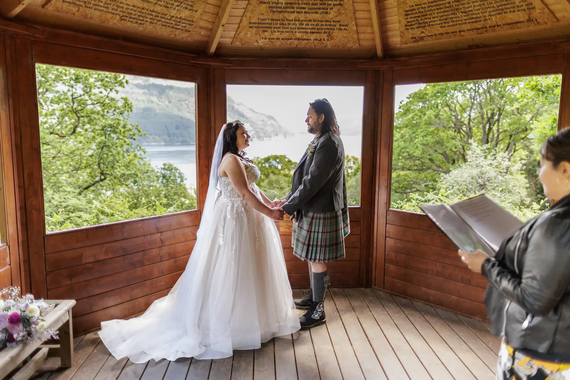 A bride in a white dress and veil holds hands with a groom in a kilt and jacket inside a wooden gazebo, surrounded by greenery and a lake view. An officiant stands to the right, holding papers and reading aloud during the ceremony.