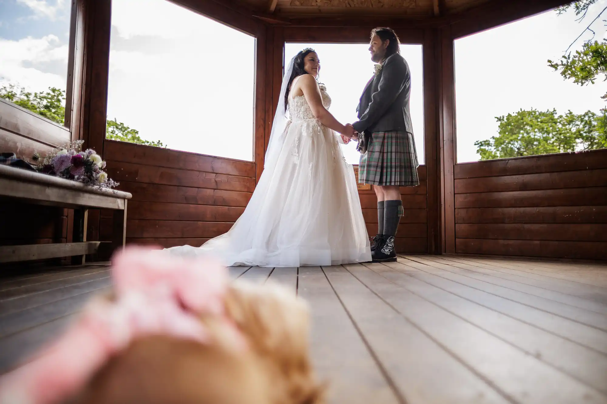 A bride in a white dress and a groom in a kilt stand holding hands in a wooden gazebo, looking at each other. In the blurred foreground, a small dog with a pink bow is partially visible, lying on the wooden floor.