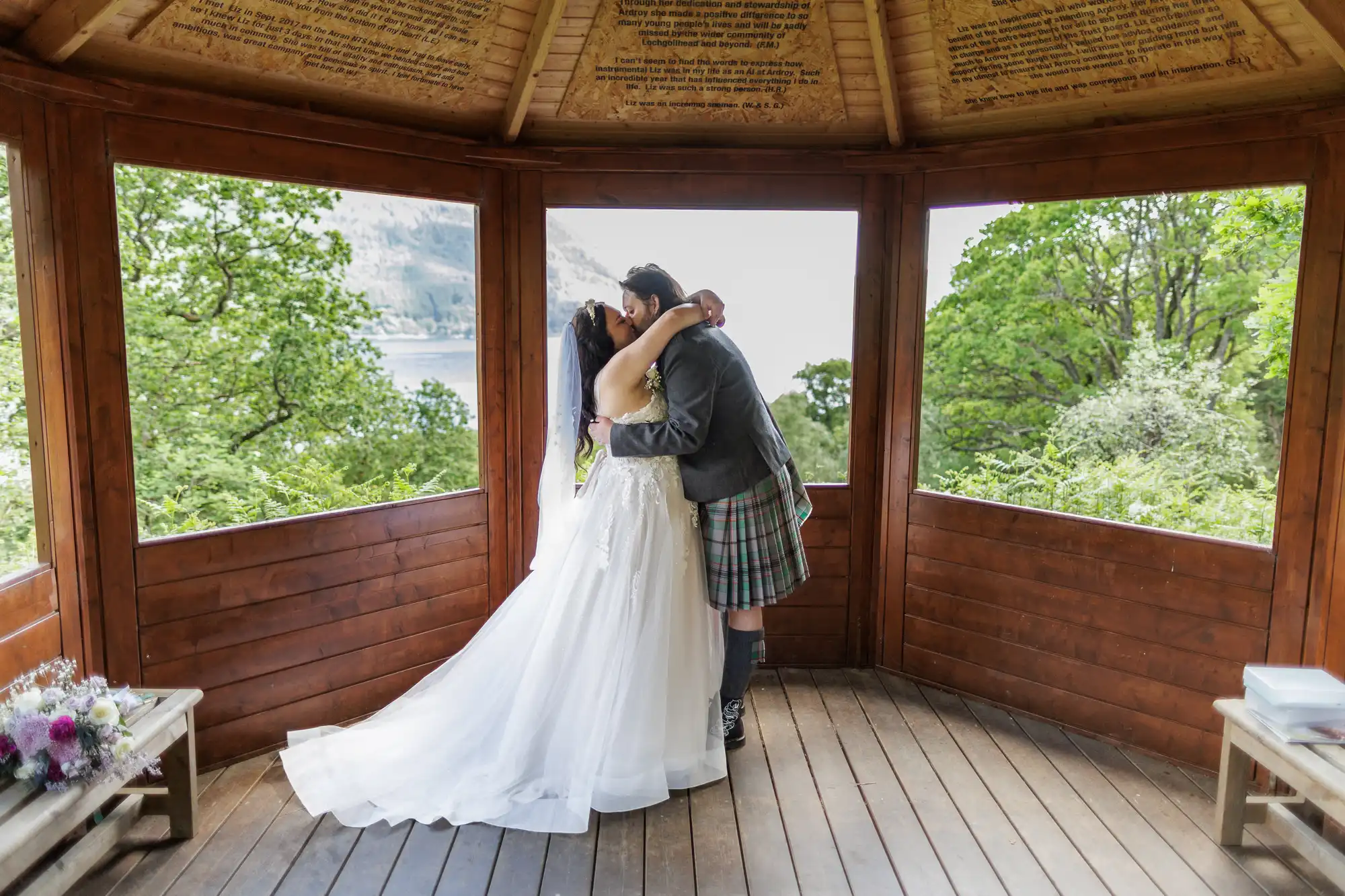 A bride in a white gown and a groom in a kilt embrace and kiss in a wooden gazebo with large windows. Lush green trees and hills are visible outside. Flowers rest on a bench, and wedding vows are inscribed on the ceiling above them.