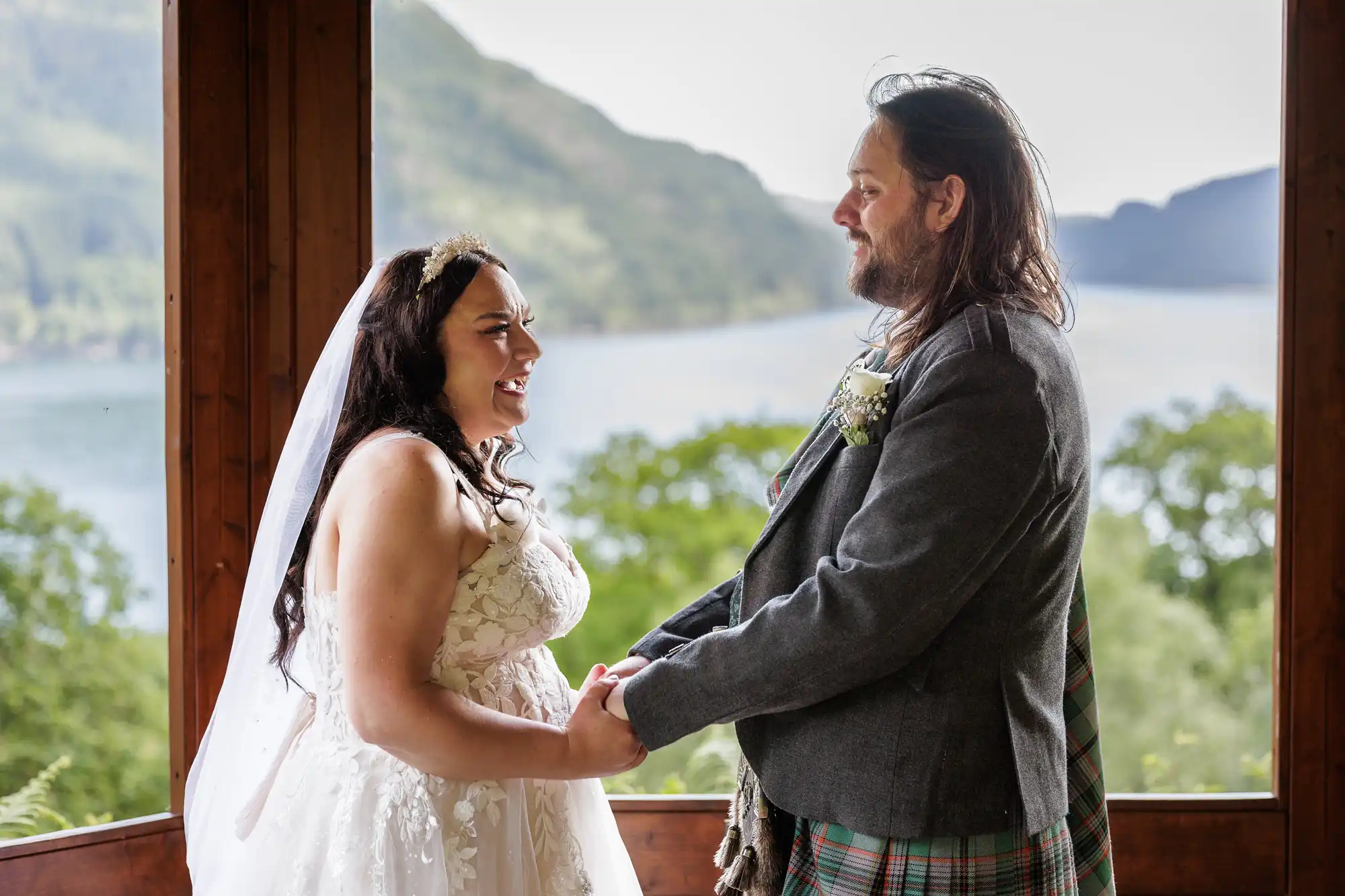Loch Goil wedding photographer photo of a bride in a white dress and veil smiles joyfully at a groom in a grey jacket and tartan kilt. They hold hands inside a wooden structure with large windows, overlooking a lush, green landscape and a lake with misty hills in the background.