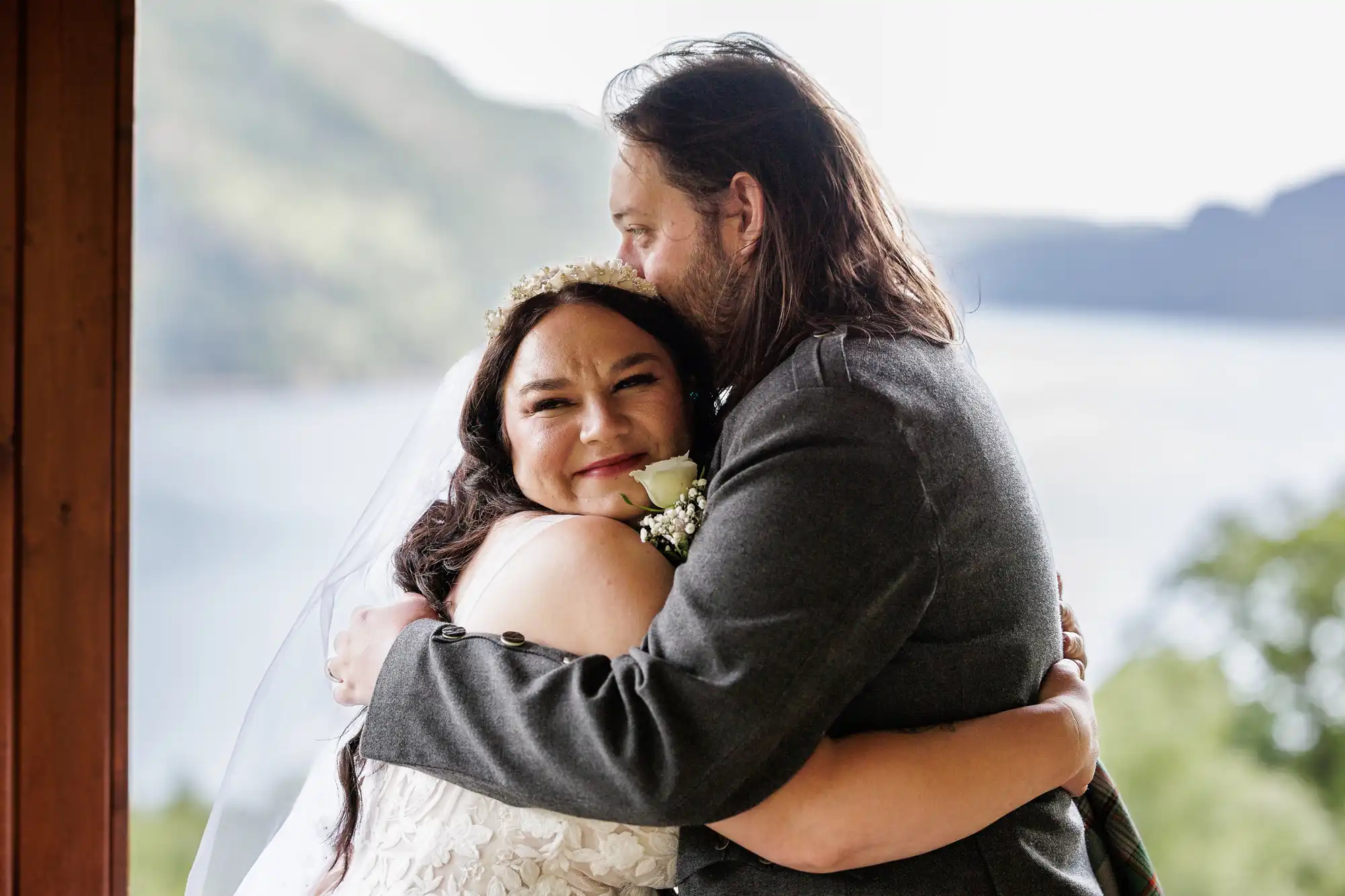 A bride in a white dress and tiara smiles as she hugs a man in a grey suit with long hair and a beard. They stand by a wooden post, with a blurred lake and mountains in the background, suggesting an outdoor wedding.