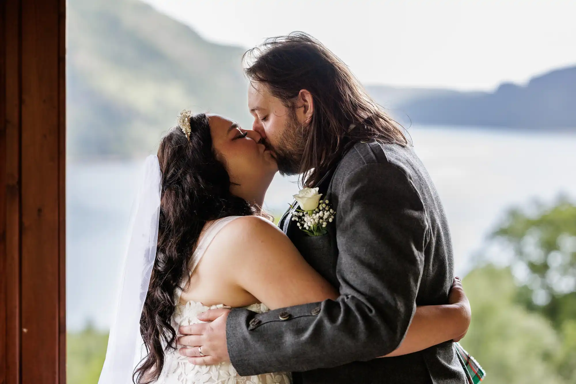 A bride and groom share a kiss on their wedding day. The bride wears a white dress and veil; the groom is in a grey suit with a buttonhole. They embrace near a wooden structure, with a scenic lake and green hills blurred in the background.