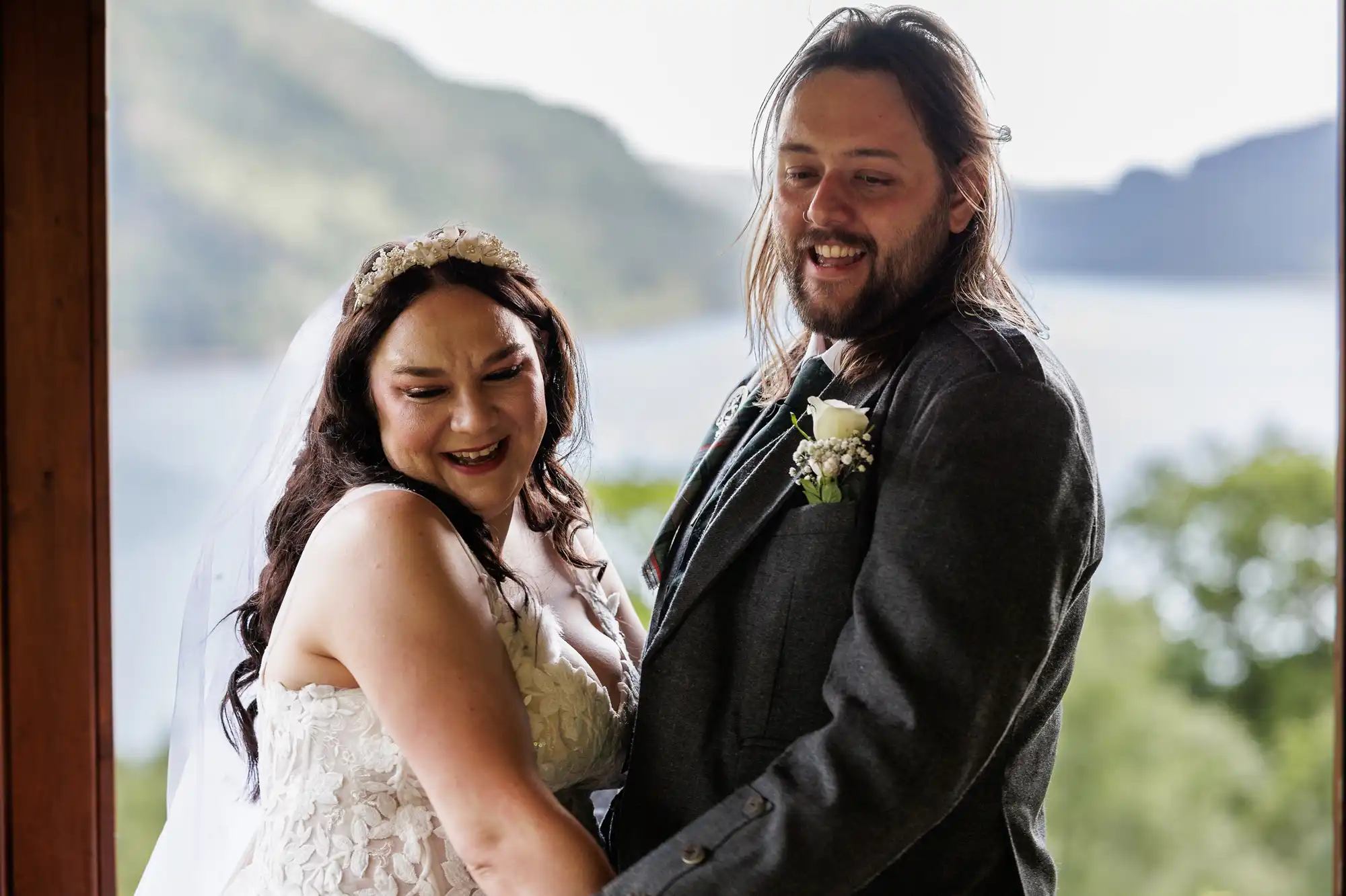 A bride in a strapless lace wedding dress and veil stands smiling next to a groom in a grey suit with a white buttonhole. They hold hands and are outdoors with blurred greenery and a lake in the background. Both look joyful.