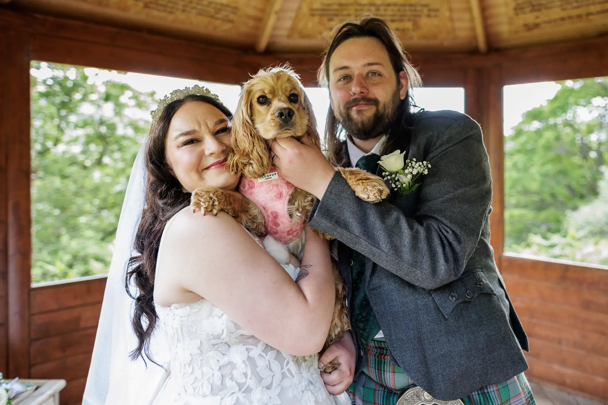 A bride in a white dress and veil and a groom in a grey jacket and tartan kilt smile whilst holding a small brown dog dressed in pink. They stand together in a wooden gazebo with greenery visible outside.