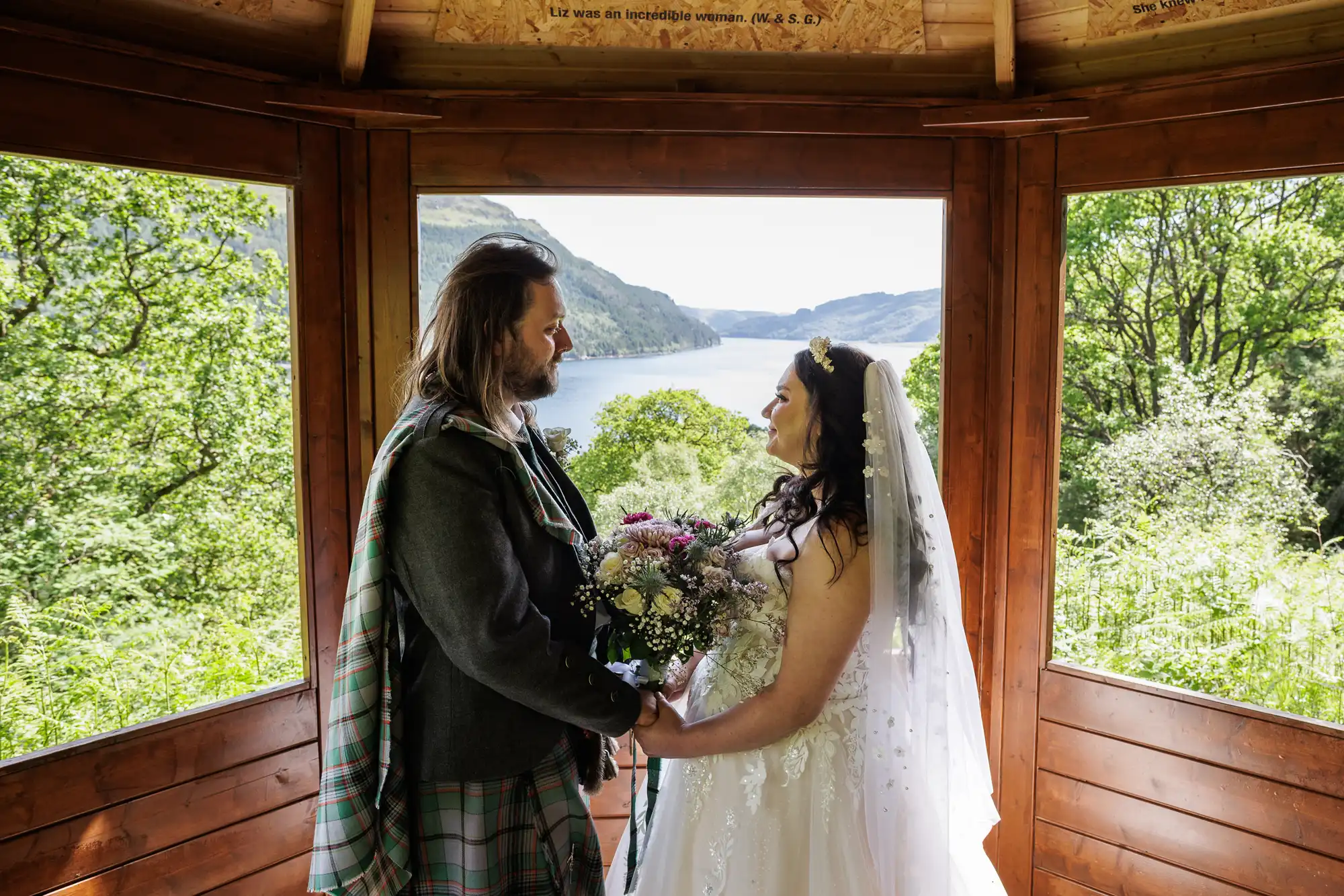 A bride in a white dress and veil holds a bouquet and faces a groom in a kilt inside a wooden gazebo. Green trees and a scenic lake stretch out through large windows behind them, with hills visible in the distance under a bright sky.