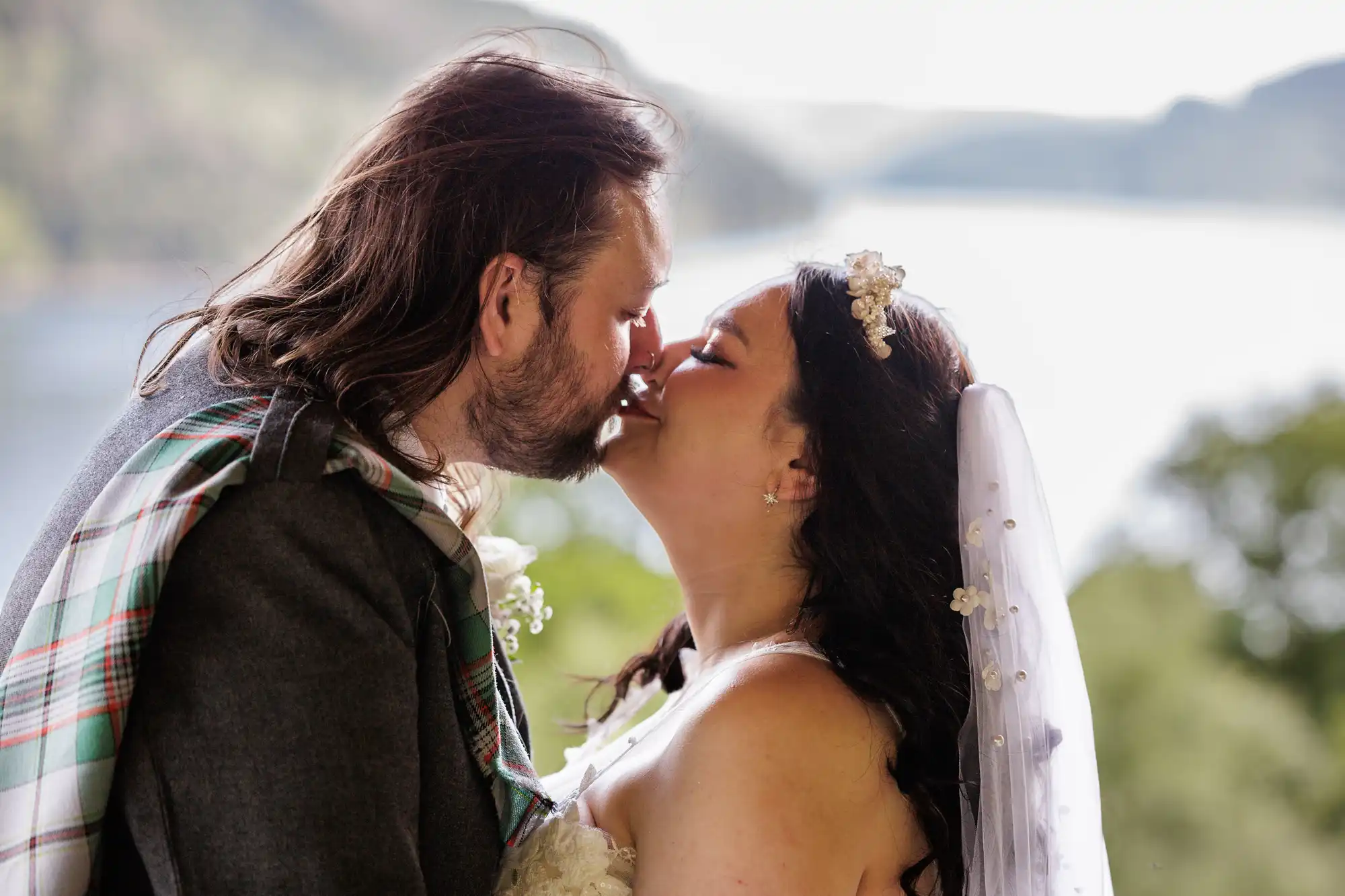 A bride and groom kiss outdoors in front of a lake and green hills. The groom wears a checked sash over a suit, and the bride wears a white dress and veil with floral decorations. Both have dark hair, and sunlight brightens the background.
