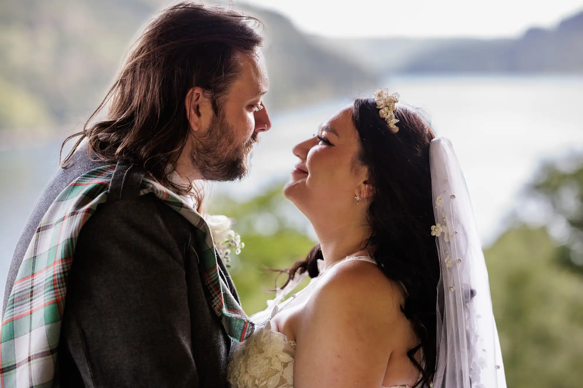 A bride and groom gaze lovingly at each other outdoors. The groom wears a grey jacket with a tartan sash, and the bride in a white dress and veil smiles up at him. A blurred, scenic landscape of trees and water is visible in the background.