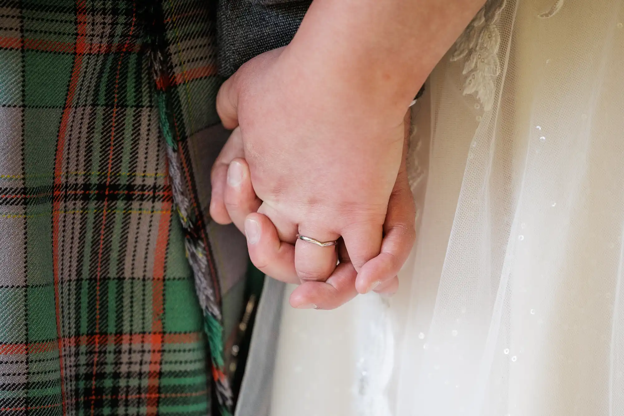 Close-up of two people holding hands; one wears a silver ring and a white lace dress, the other wears a green checked garment, possibly a kilt. The image captures tenderness, with focus on their intertwined fingers and contrasting fabrics.