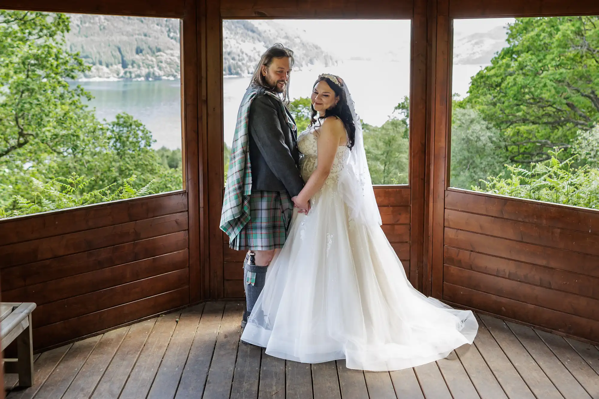 A couple stands hand in hand in a wooden gazebo, dressed in wedding attire. The groom wears a traditional kilt, and the bride has a flowing white gown and veil. Green trees and a lake are visible in the scenic background. Both look towards the camera.