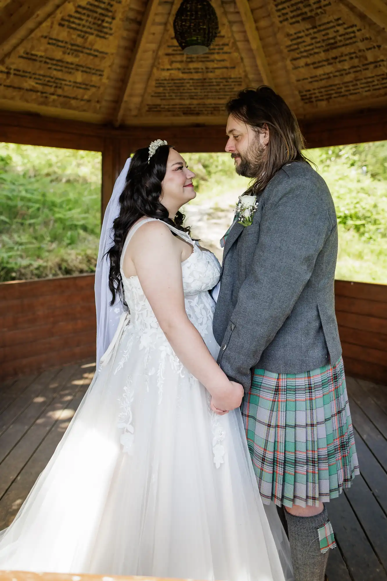 A bride in a white gown and veil holds hands with a groom in a grey jacket and tartan kilt. They face each other, smiling, inside a wooden gazebo with sunlight and greenery visible in the background.