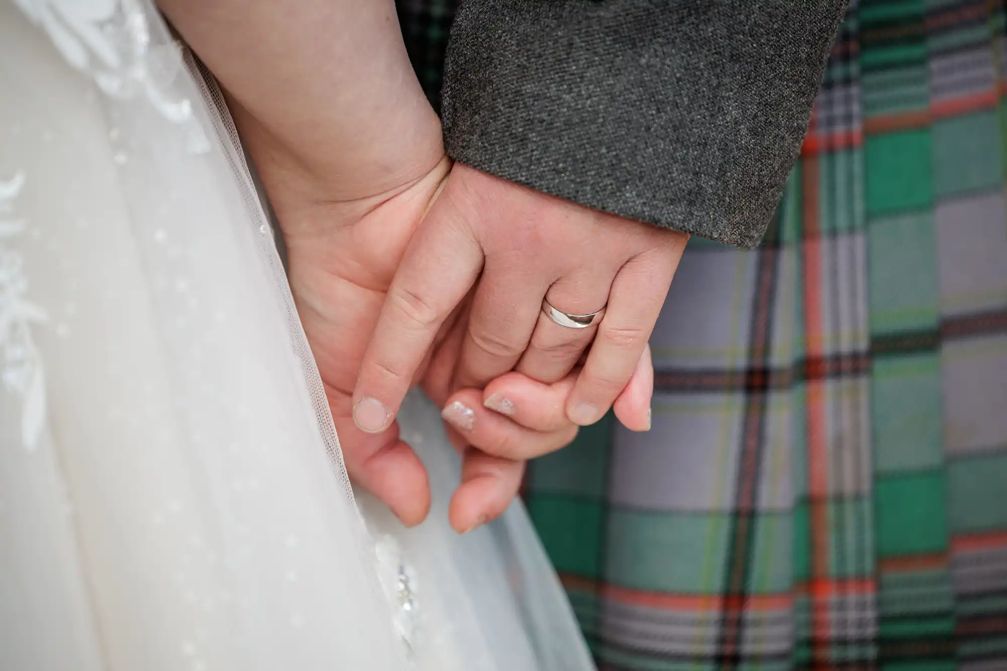 Close-up of two people holding hands; one wears a wedding ring and a white lace dress, and the other wears a grey jacket and tartan plaid fabric, suggesting a wedding or formal event.
