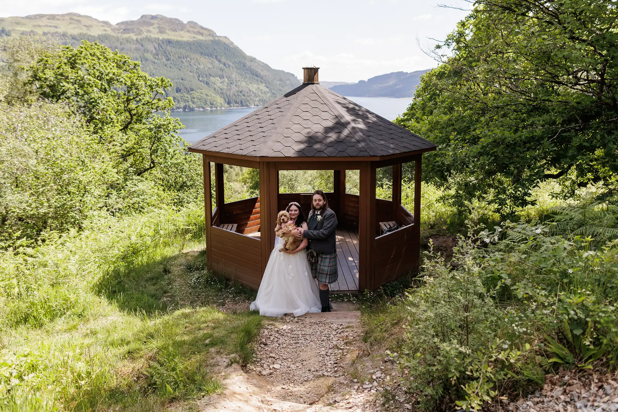 A bride in a white gown and a groom in a kilt stand smiling, holding a bouquet, in front of a wooden gazebo surrounded by lush greenery. Behind them, a lake and forested mountains are visible under a partly cloudy sky.