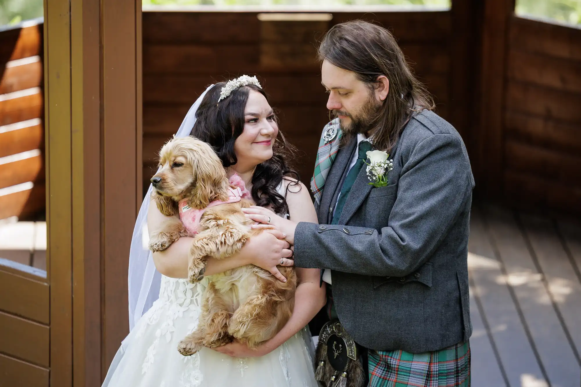 A bride in a white gown and veil smiles while holding a brown dog with a pink scarf. A groom in a grey jacket and checked kilt stands beside her, gently touching the dog. They stand on a wooden porch, surrounded by natural light.