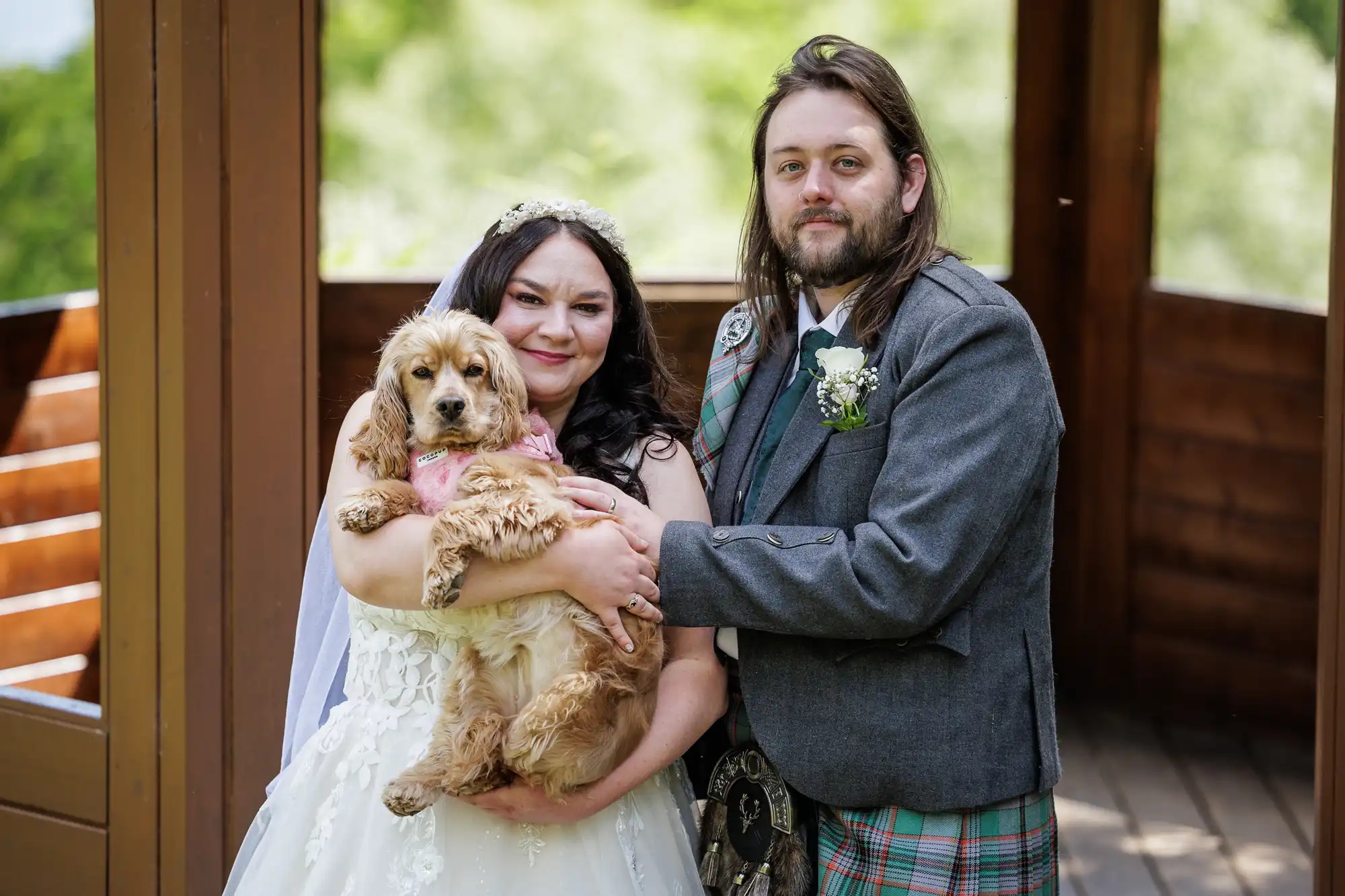 A bride in a white dress and veil and a groom in a grey suit with tartan accents stand together outside, smiling and holding a tan cocker spaniel dog. The background is green and wooden, suggesting a natural, outdoor setting.