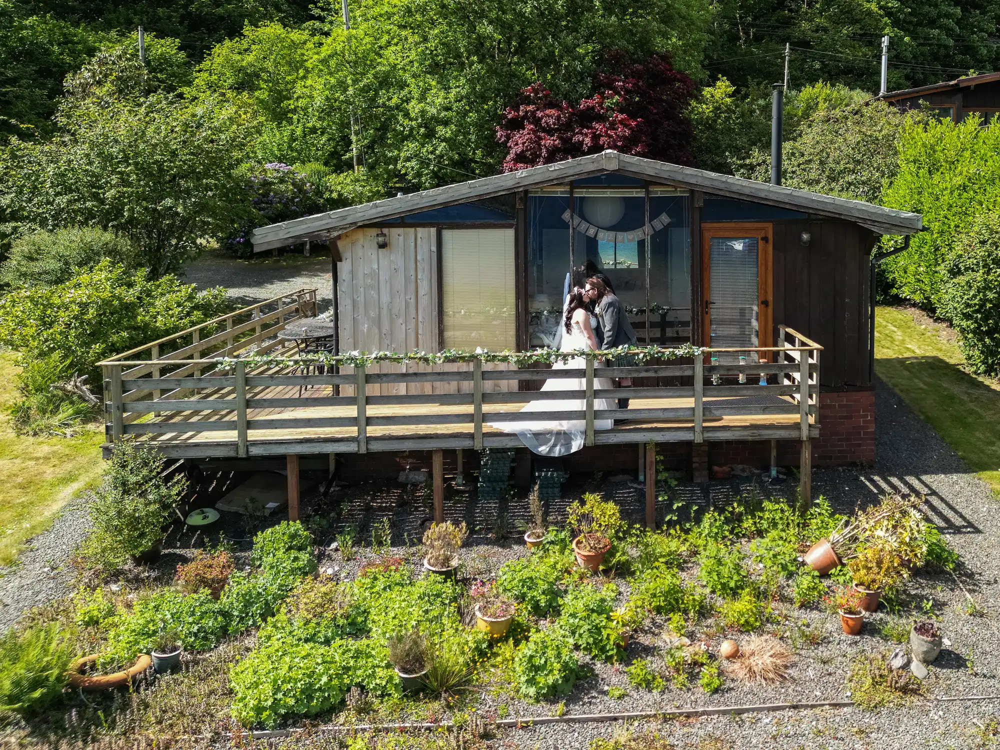 A bride and groom embrace and kiss on the wooden deck of a rustic cabin surrounded by lush greenery, potted plants, and garden beds on a sunny day. The cabin has large windows and is nestled among trees and shrubs.