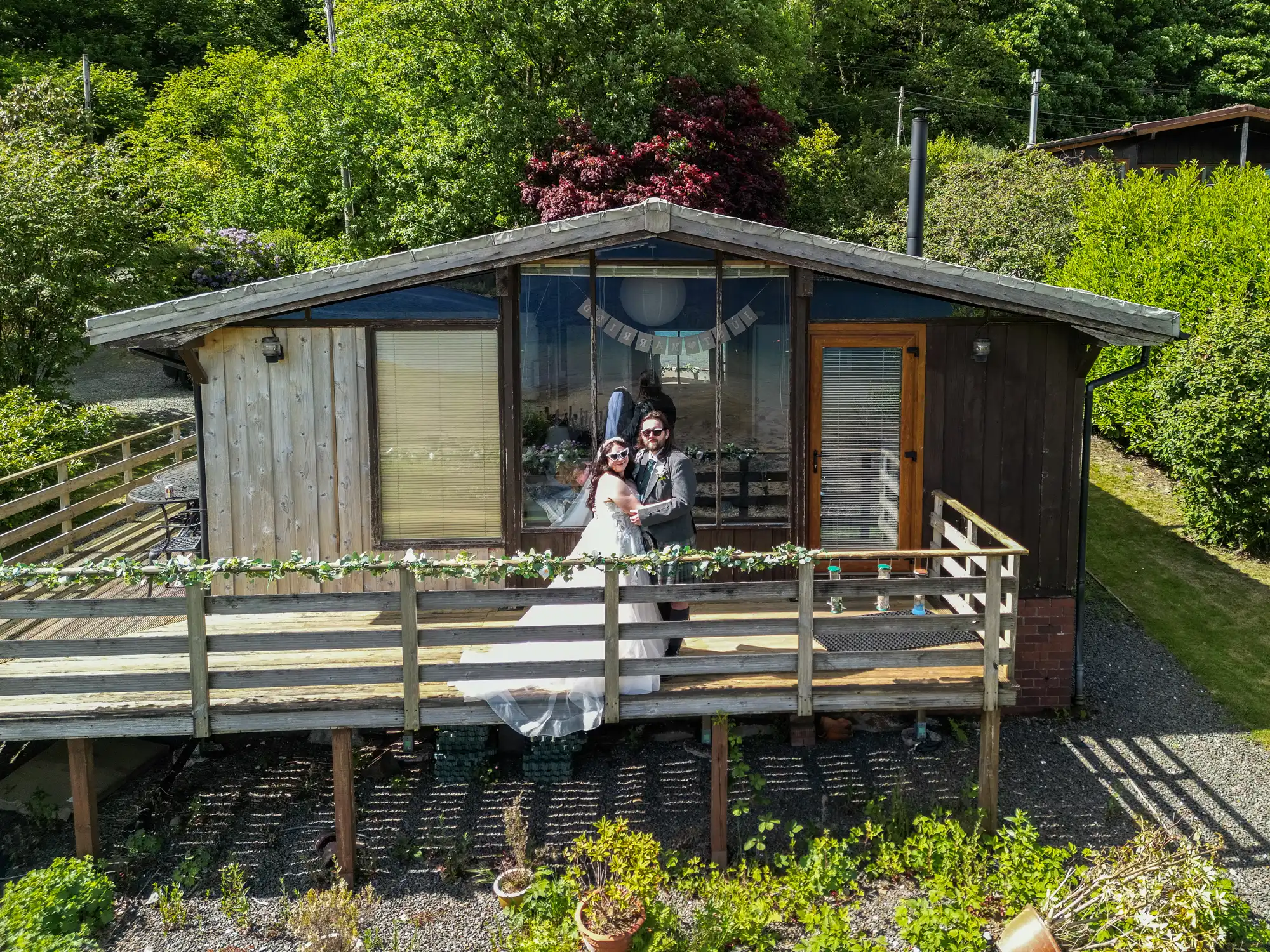A bride and groom, both wearing sunglasses, stand on the wooden decking of a rustic cabin surrounded by green trees and bushes. The bride wears a white dress and veil, the groom a grey suit. A banner with bunting decorates the front of the cabin.