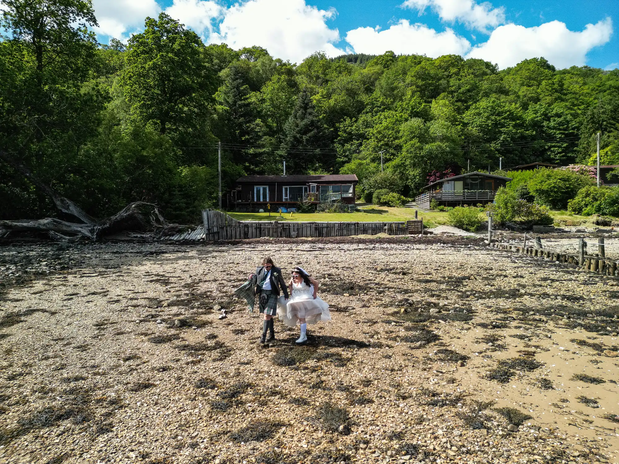 A bride in a white dress and a groom in a dark suit joyfully hold hands whilst walking on a rocky, sandy shore. Behind them are green trees and two wooden houses under a bright blue sky with scattered clouds.