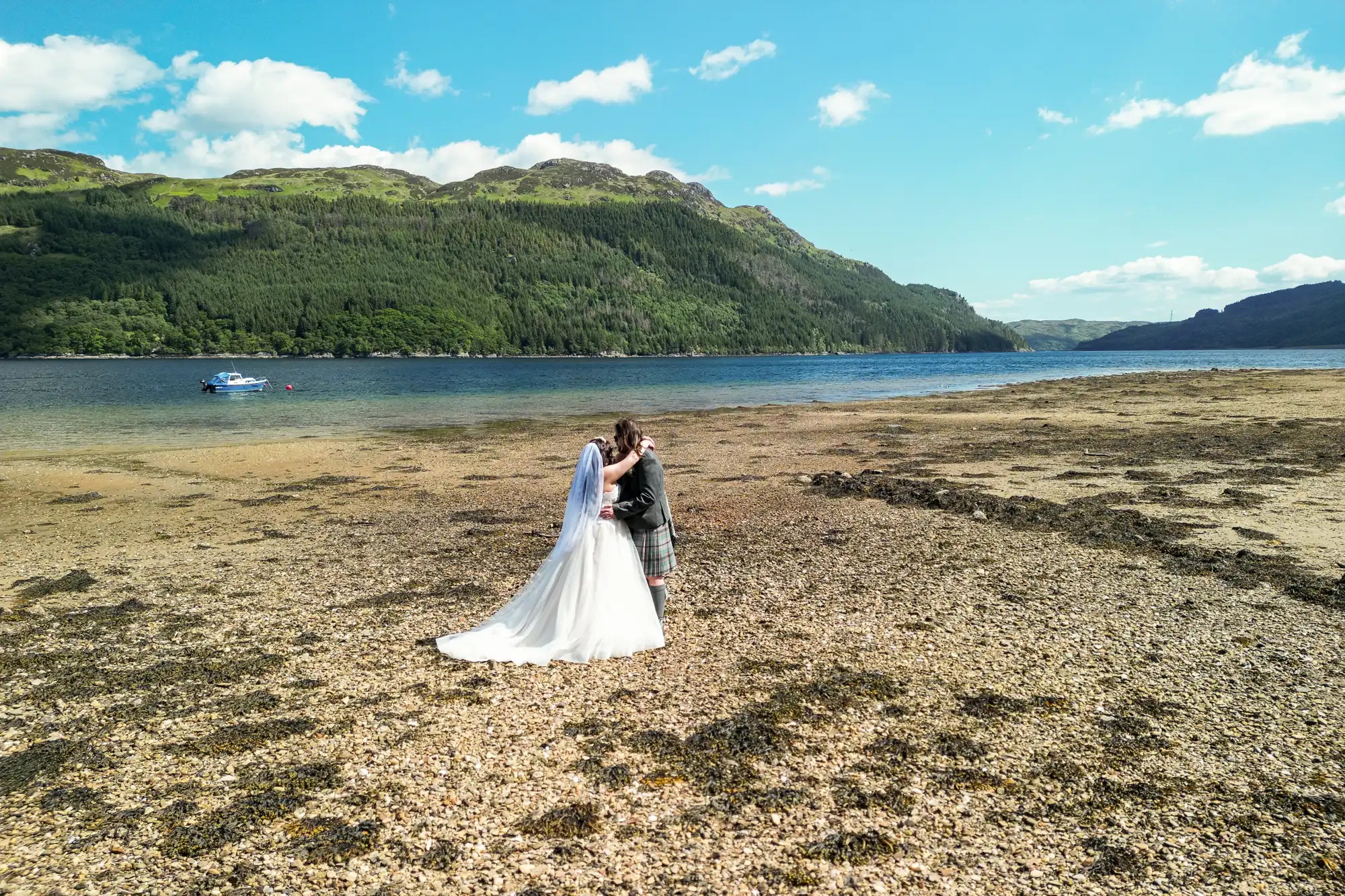 A bride in a white gown and veil kisses a groom in a traditional kilt on a rocky shore beside a calm lake, with forested hills and a blue sky with scattered clouds in the background. A small boat floats on the water in the distance.