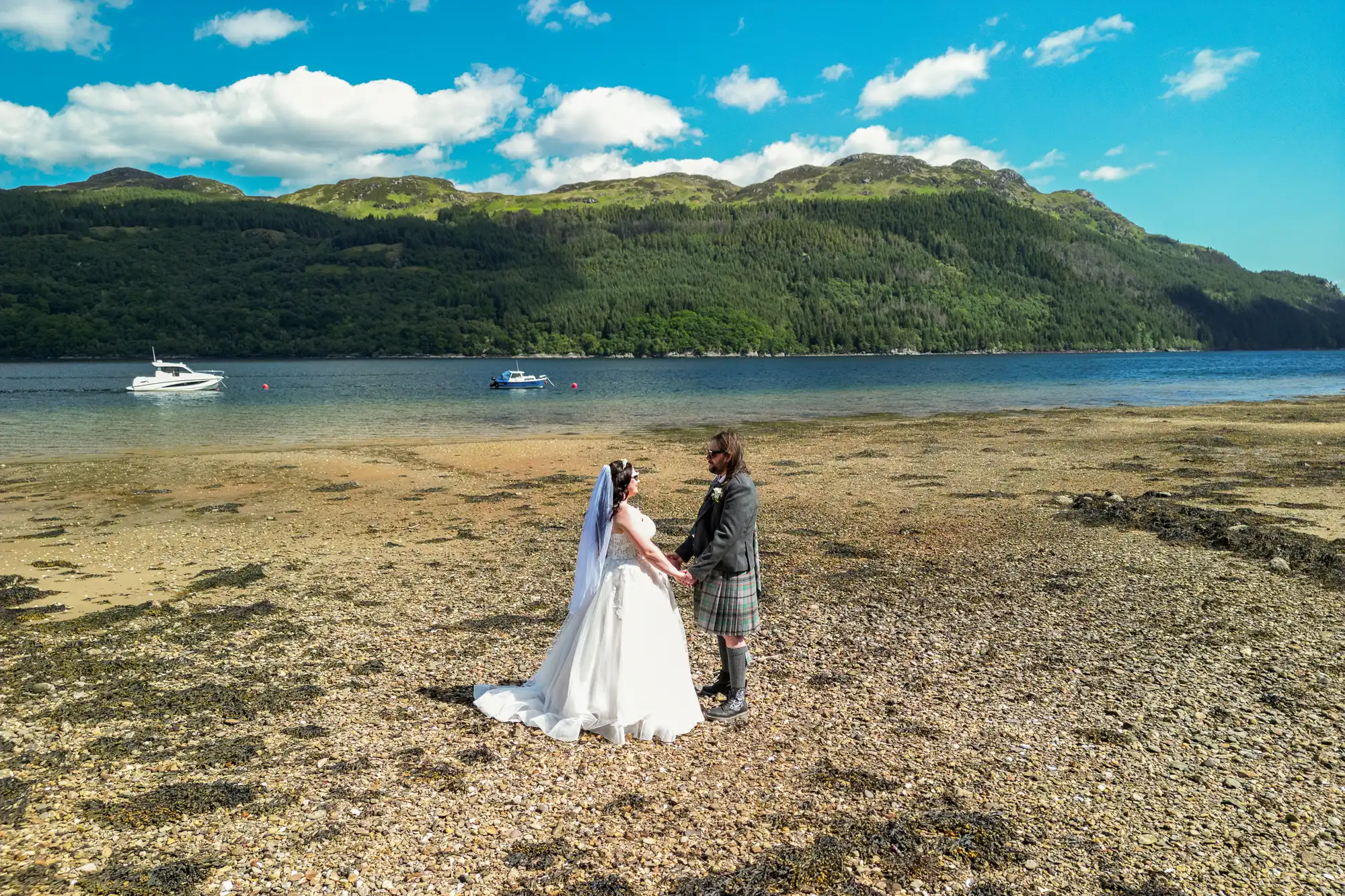 A bride in a white dress and a groom in a kilt stand holding hands on a rocky shore by a calm lake, with green hills and mountains in the background under a bright blue sky with scattered clouds. Two boats float on the water.