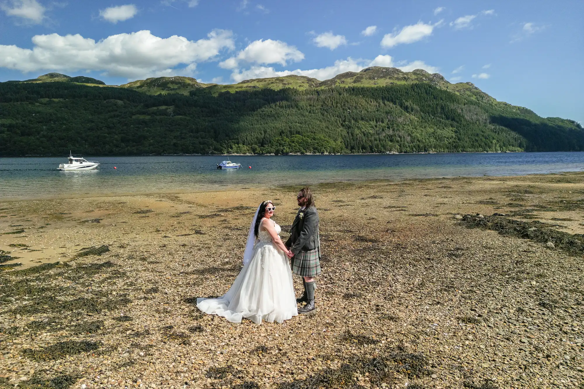 A bride in a white dress and a groom in a kilt hold hands on a rocky shore, facing each other, with a lake and forested hills in the background. Two boats float on the water under a partly cloudy blue sky.