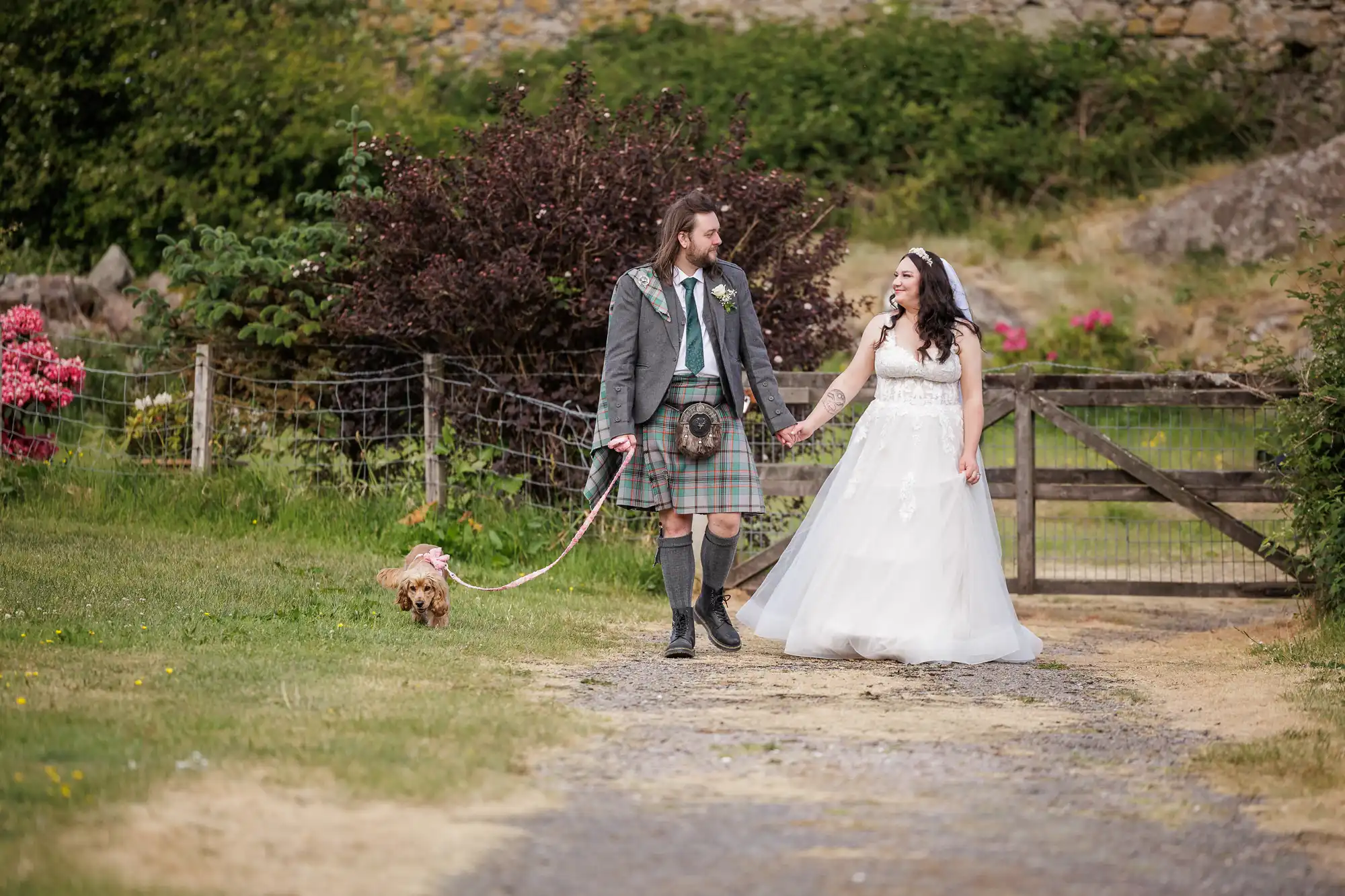 A couple in wedding attire—man in a kilt and jacket, woman in a white gown—walk hand in hand outdoors on a gravel path. The man holds a dog’s lead. There’s greenery, a wooden gate, and shrubs with pink flowers in the background.