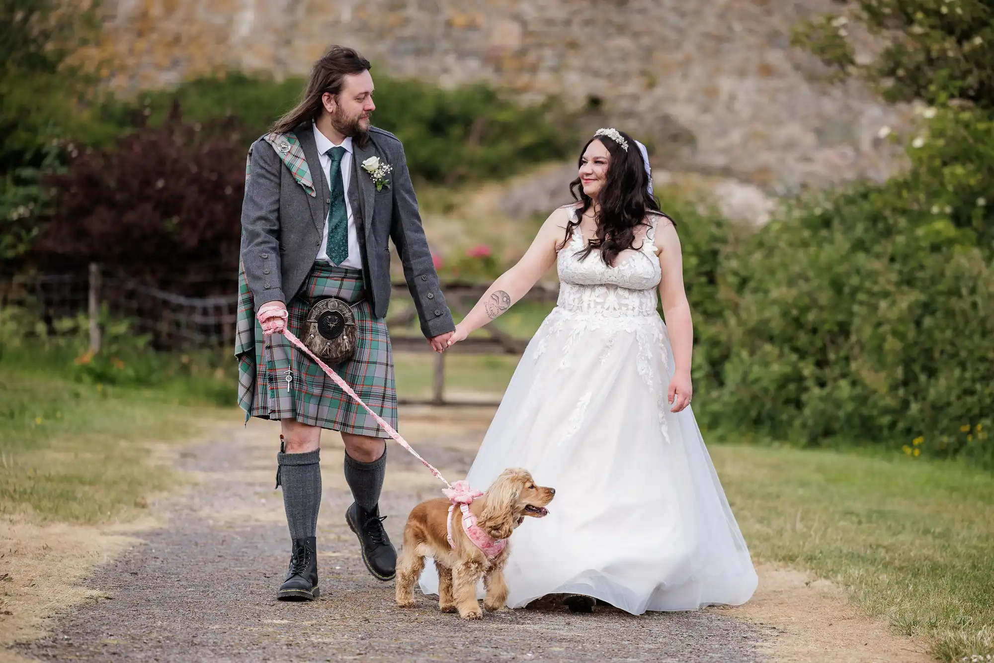 A bride in a white dress and a groom in a kilt walk hand in hand outdoors, smiling at each other. They are walking a small brown dog on a pink lead. The background is grassy with bushes and a stone wall.