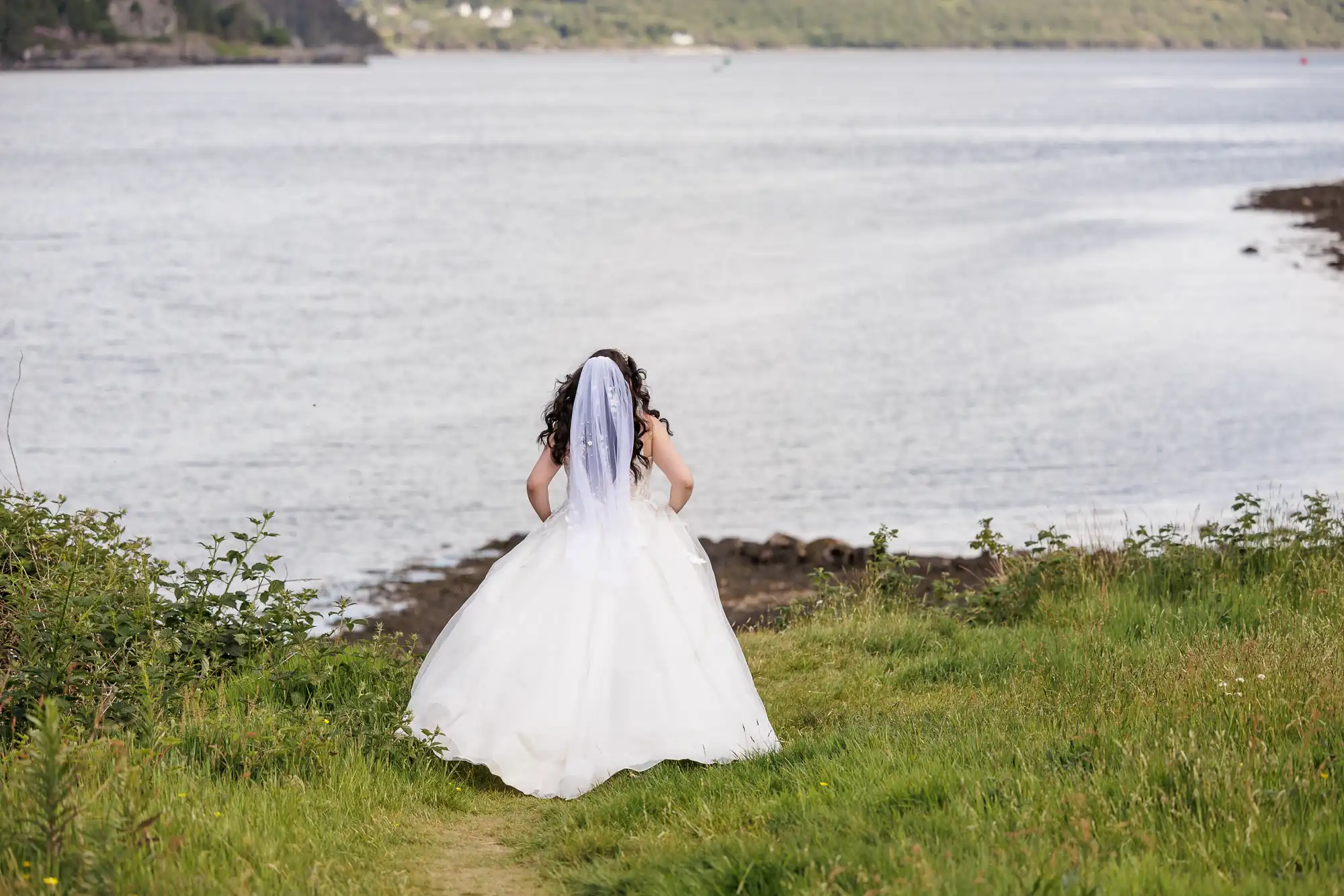 A bride in a white wedding dress and veil stands on grass, facing a calm body of water. Her back is to the camera, and she has long dark hair. The scene is peaceful, with greenery and distant hills across the shore.