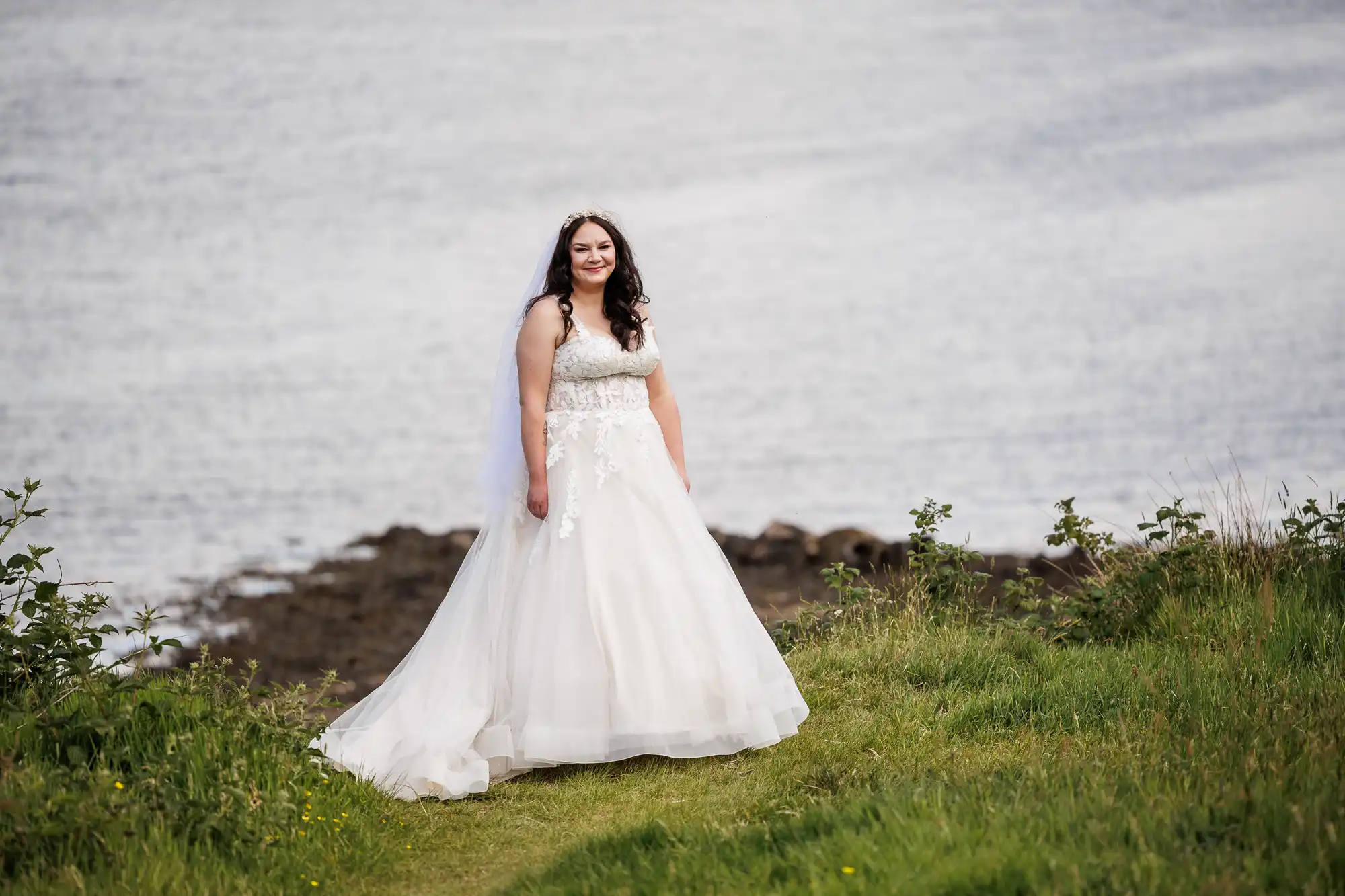 A woman in a white lace wedding dress stands on green grass near a rocky shoreline, smiling at the camera. Her dark hair is loose with a veil, and the calm water behind her stretches across the background. It’s a bright, outdoor setting.