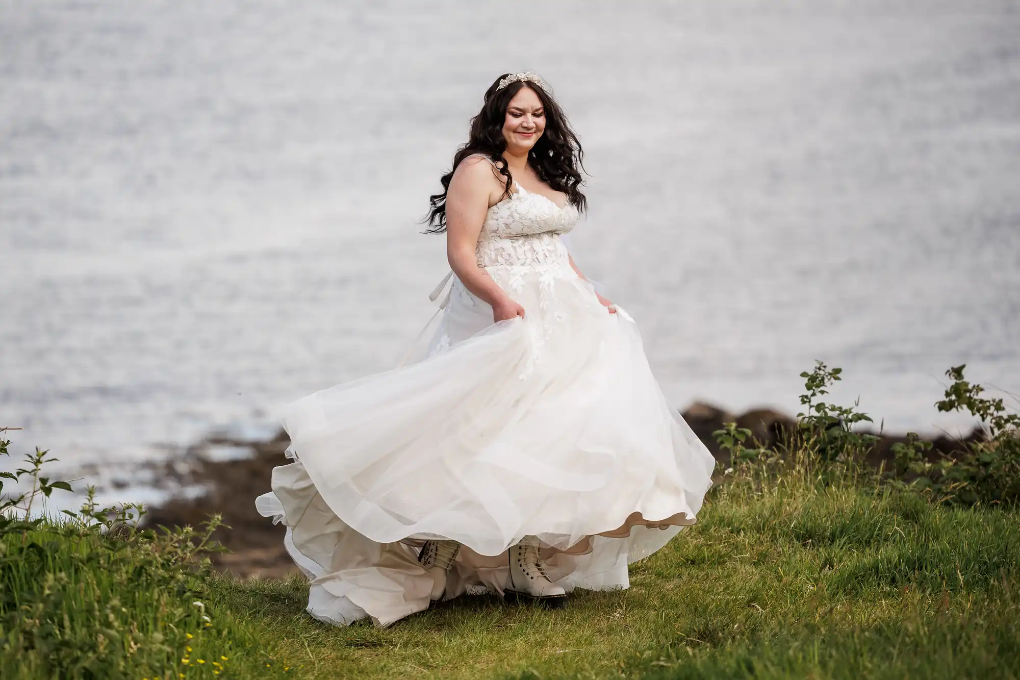 A woman with long dark hair in a white wedding dress smiles as she twirls outdoors. The flowing gown billows around her. She stands on green grass beside water, with small plants and a rocky shore in the background.