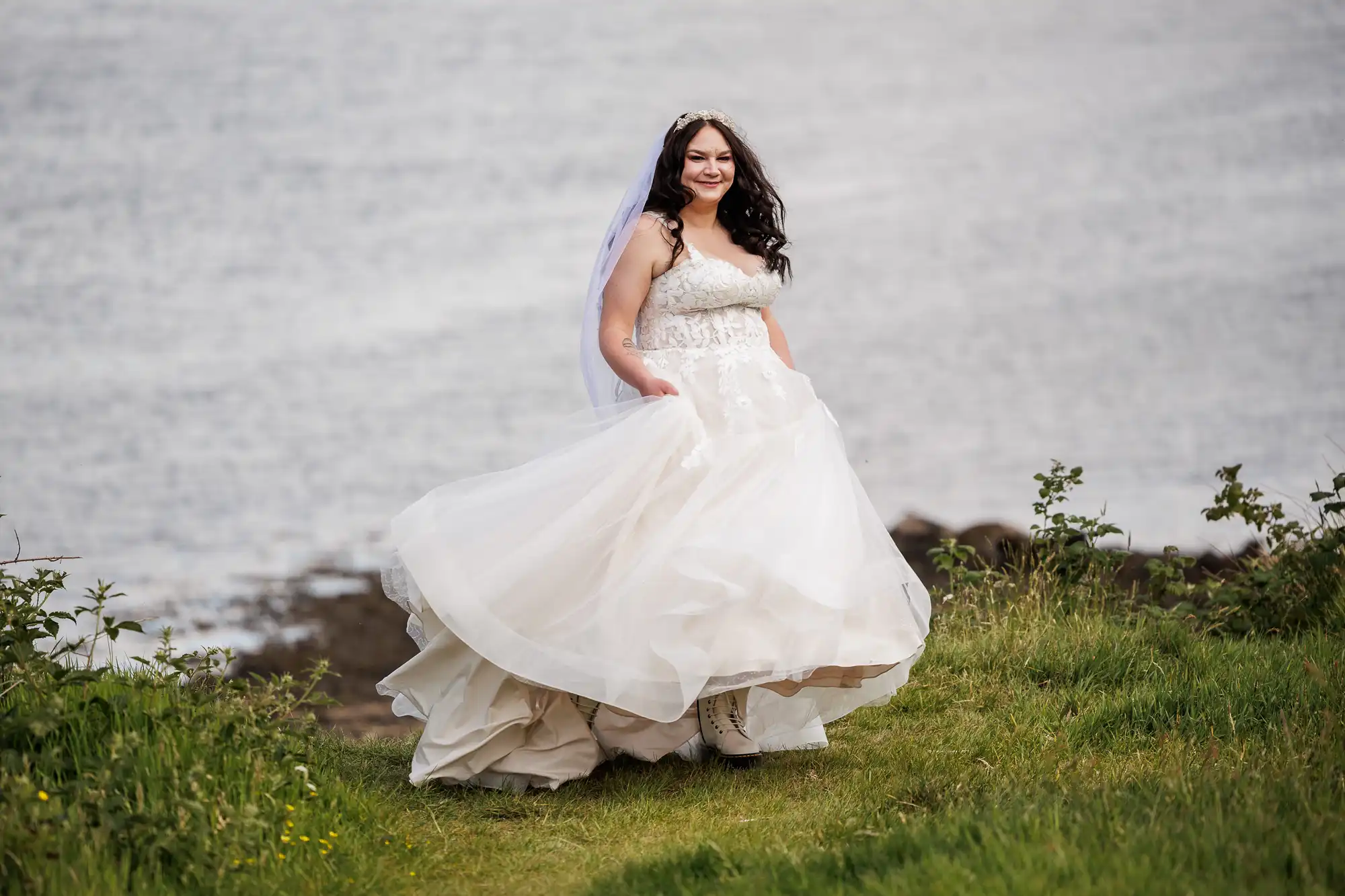 A smiling bride in a white wedding dress and veil stands on green grass near a body of water. She lifts up her dress, revealing white boots underneath. Bushes and wildflowers are visible around her. The background shows water with gentle ripples.