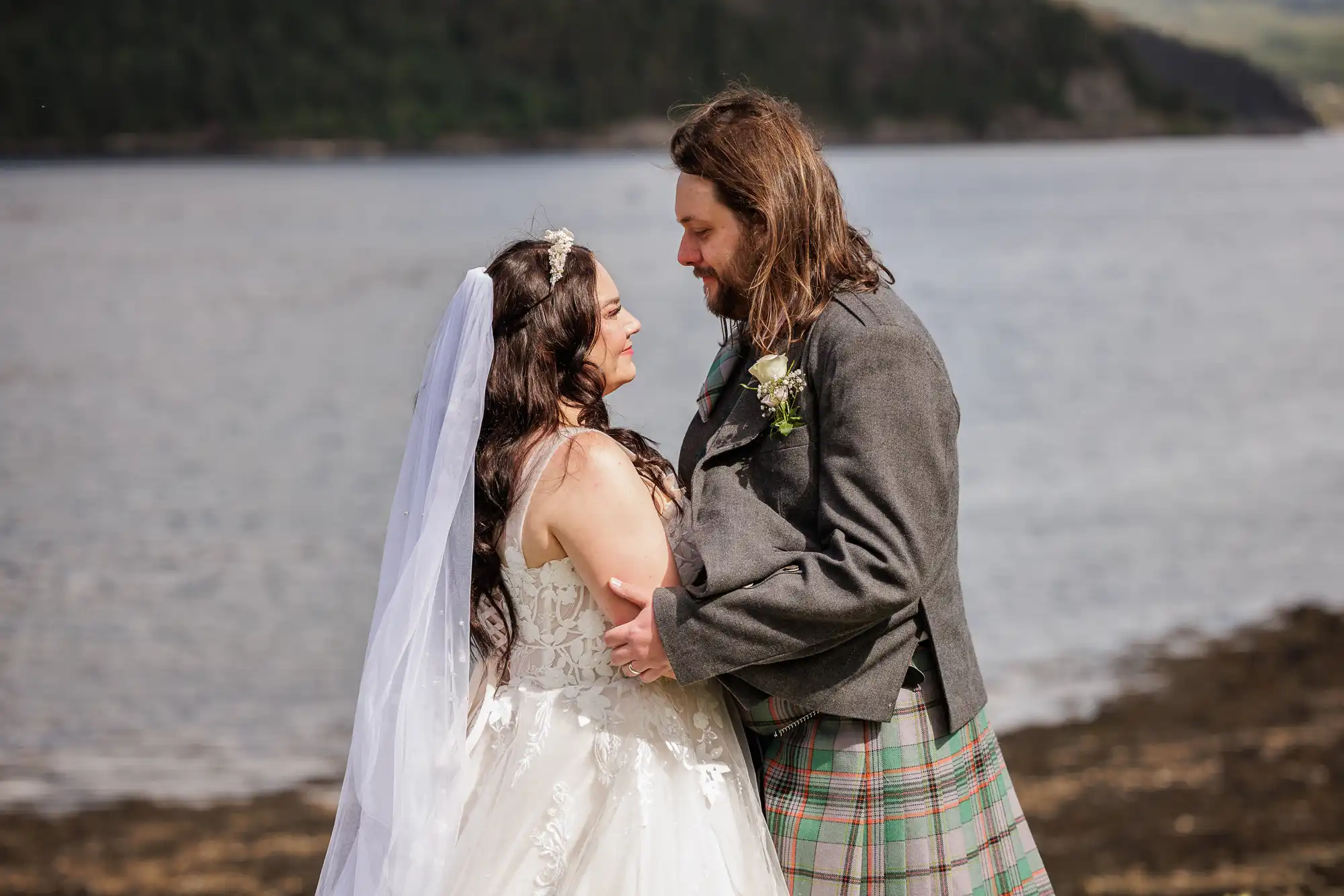 A bride in a white dress and veil faces a groom in a grey jacket and tartan kilt by a lakeside, holding each other and gazing into each other's eyes with forested hills and water in the background.
