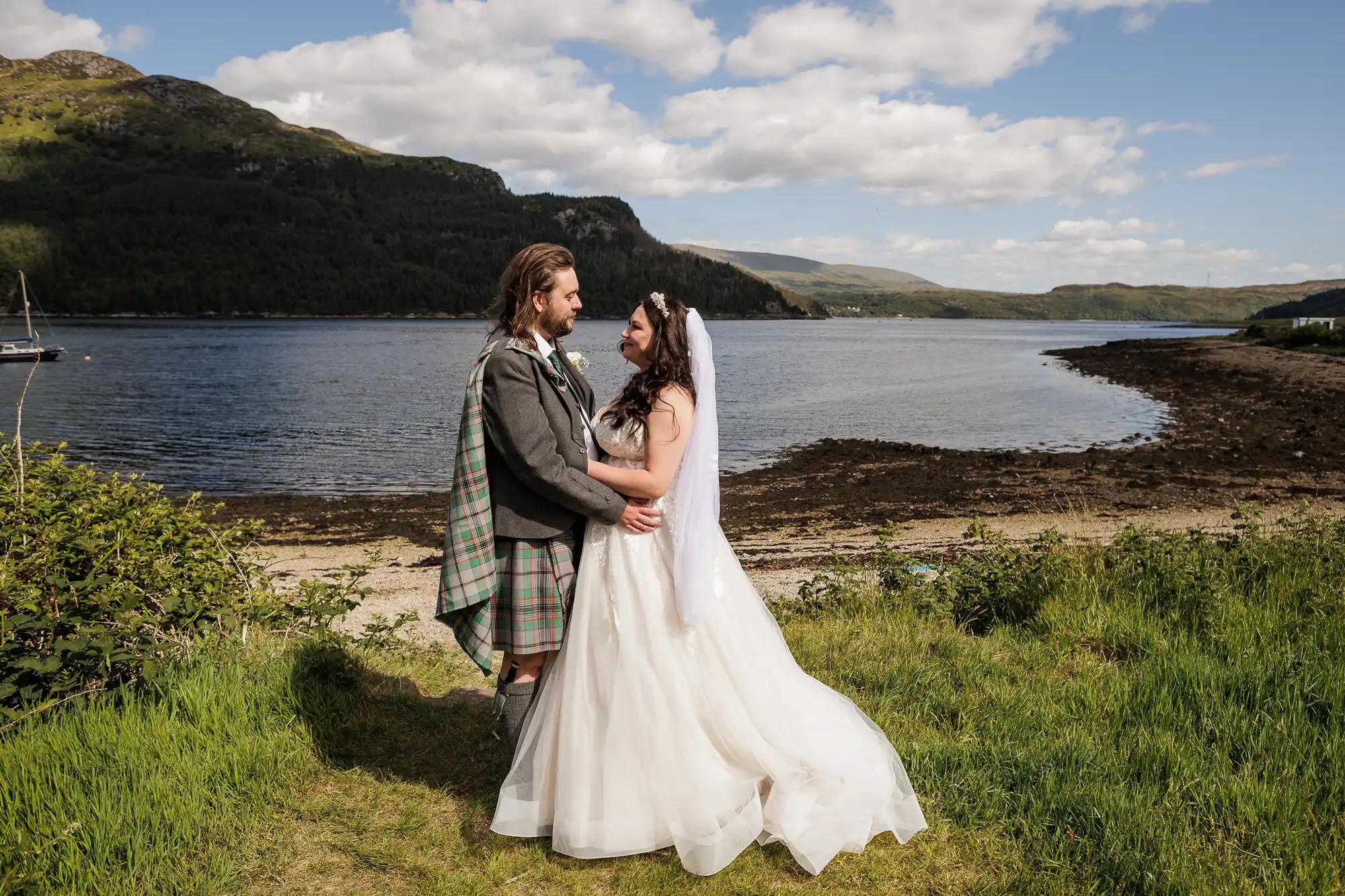 A bride in a white dress and veil and a groom in a kilt and jacket stand embracing on grass beside a lake, with mountains and a partly cloudy sky in the background. The scene is bright and peaceful, evoking a romantic outdoor wedding.