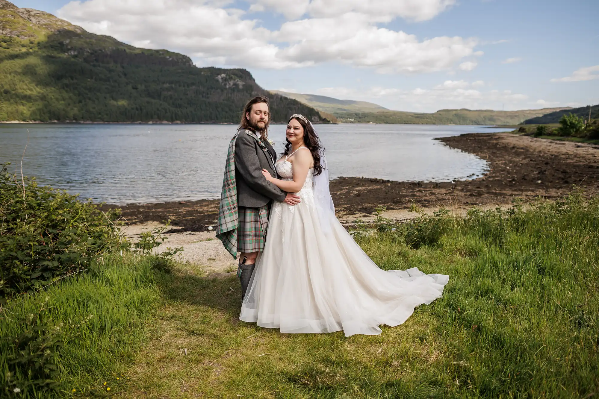 Loch Goil wedding photographer photo of a bride in a white gown and a groom in a grey traditional Scottish kilt embrace on grassy land by a lake, with green hills, a partly cloudy blue sky, and mountains in the background.