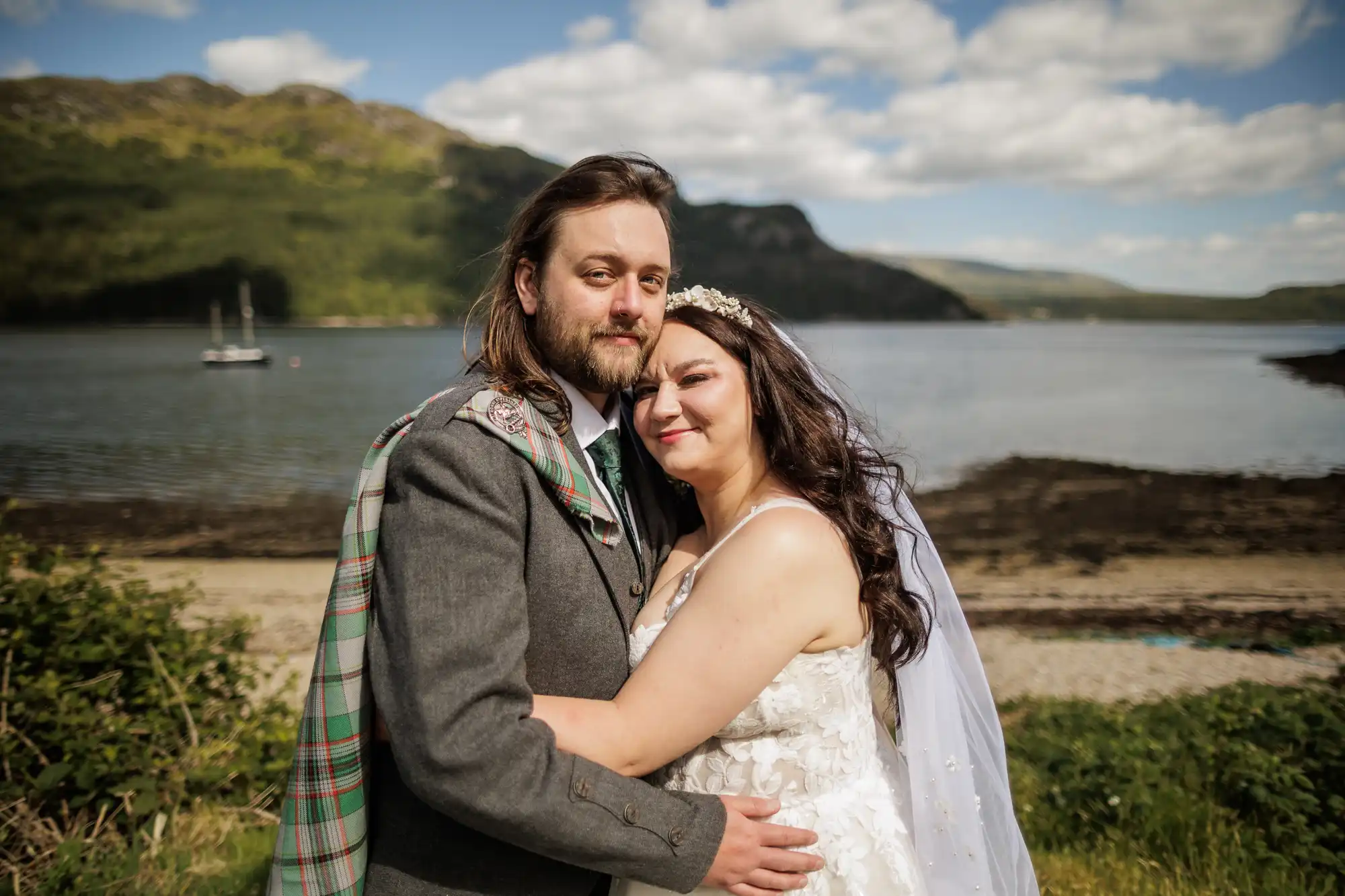 A couple in wedding attire embraces outdoors by a scenic lake. The groom wears a grey suit with a tartan sash, and the bride wears a white lace dress with a veil and headpiece. Green hills and a partly cloudy sky appear in the background.