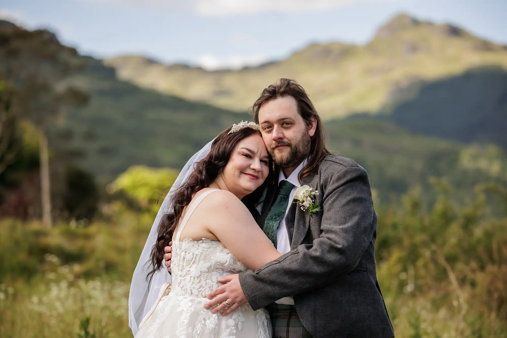 A bride and groom embrace outdoors, smiling at the camera. The bride wears a white dress and veil, while the groom is in a grey suit with a buttonhole. Lush green hills and mountains are in the background under a blue sky.