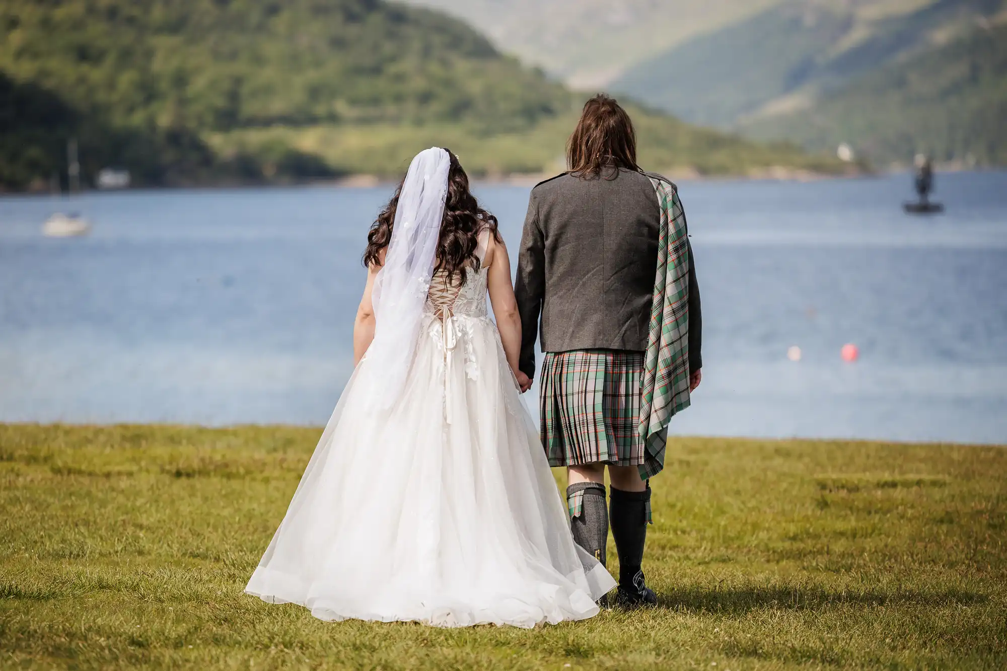 A bride in a white dress and veil stands hand in hand with a groom in a kilt and tartan sash, both facing a scenic lake surrounded by green hills and trees, with boats visible on the water in the background.