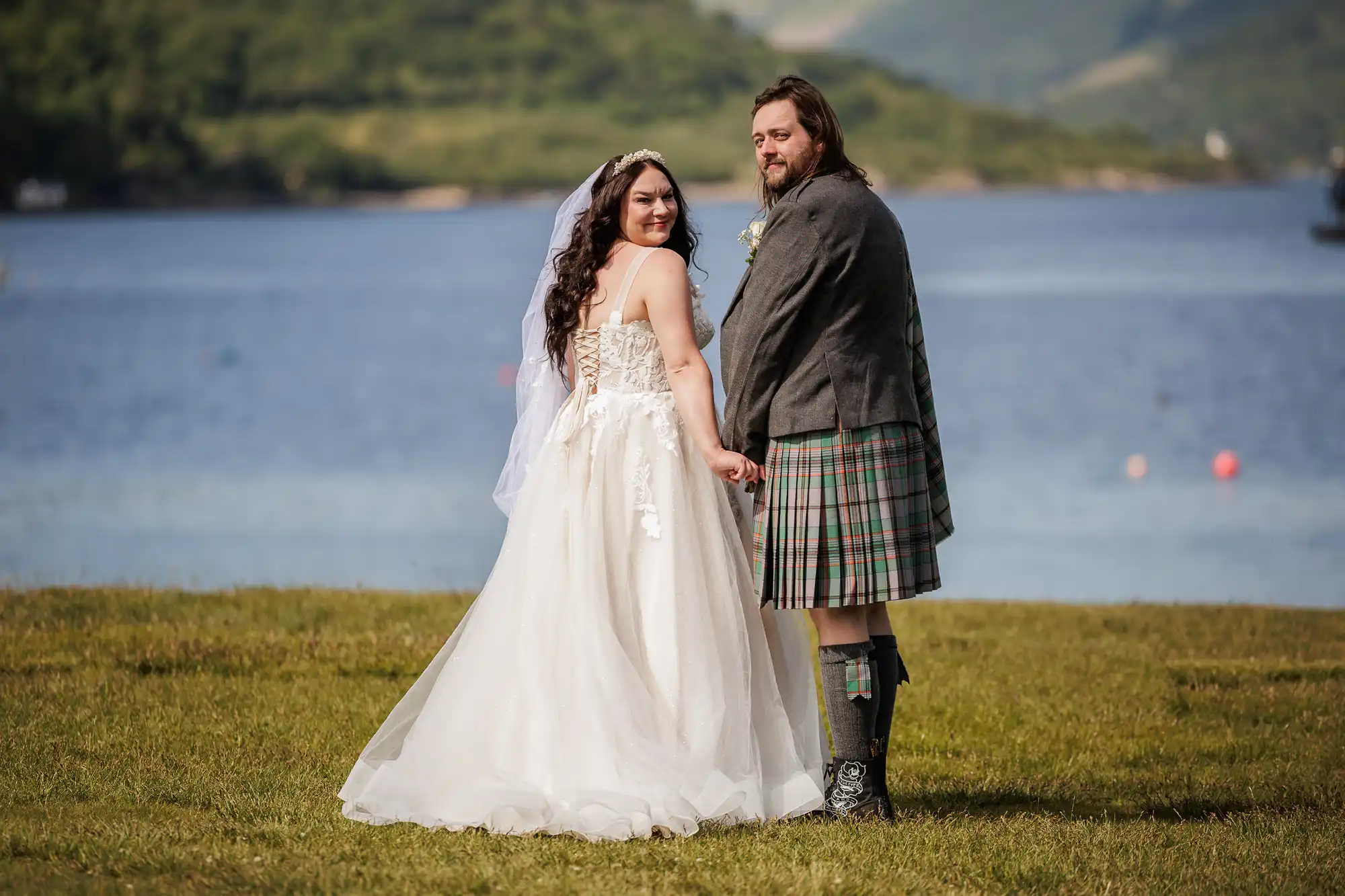 A bride in a white gown and veil stands holding hands with a groom in a tartan kilt and grey jacket. They face away but look back at the camera, standing on grass by a blue lake with green hills in the background, on a sunny day.