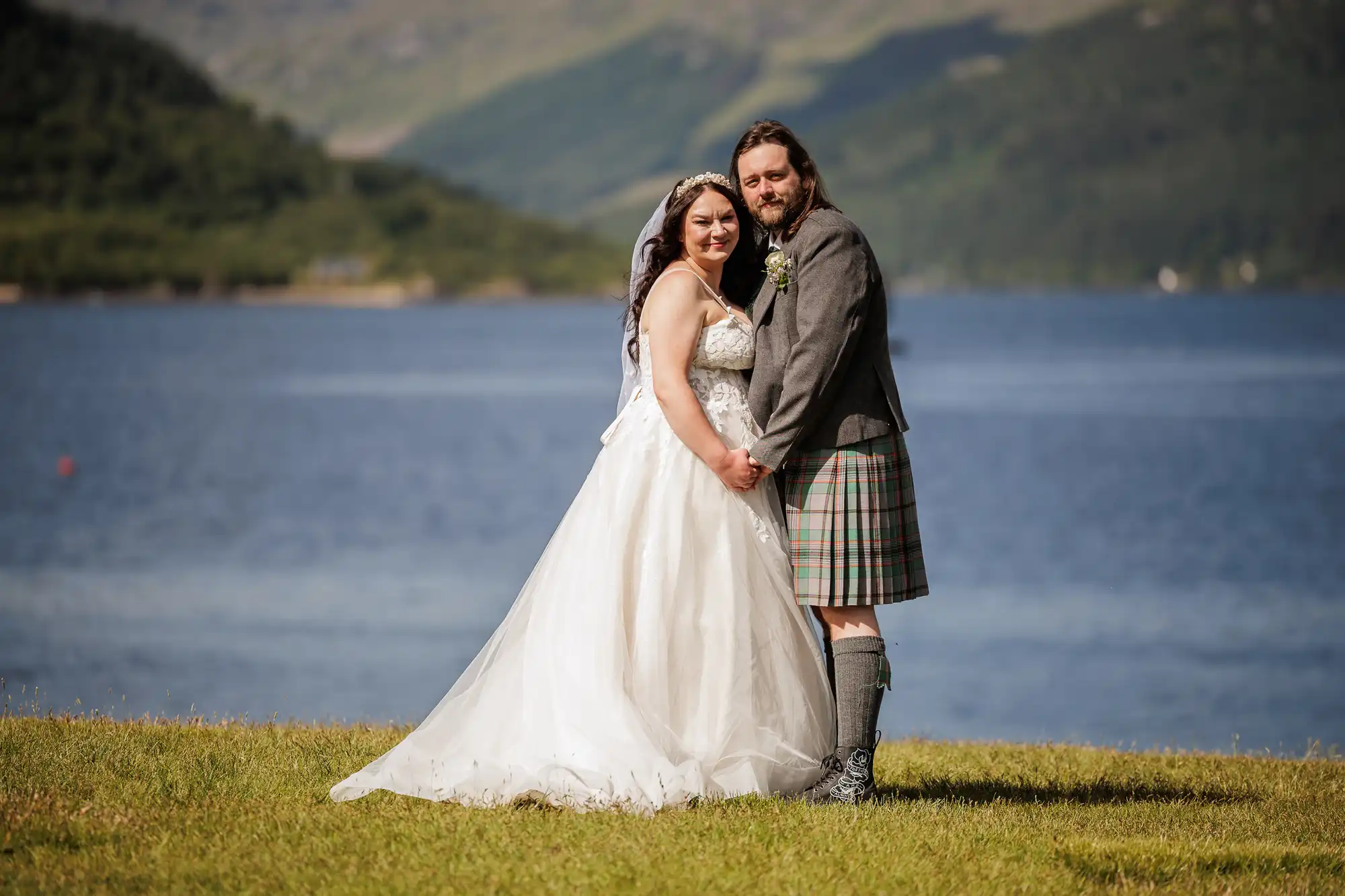 A bride in a white gown and a groom in a grey jacket with a Scottish kilt stand hand in hand on grass by a blue lake, with green hills and mountains in the background under a sunny sky.
