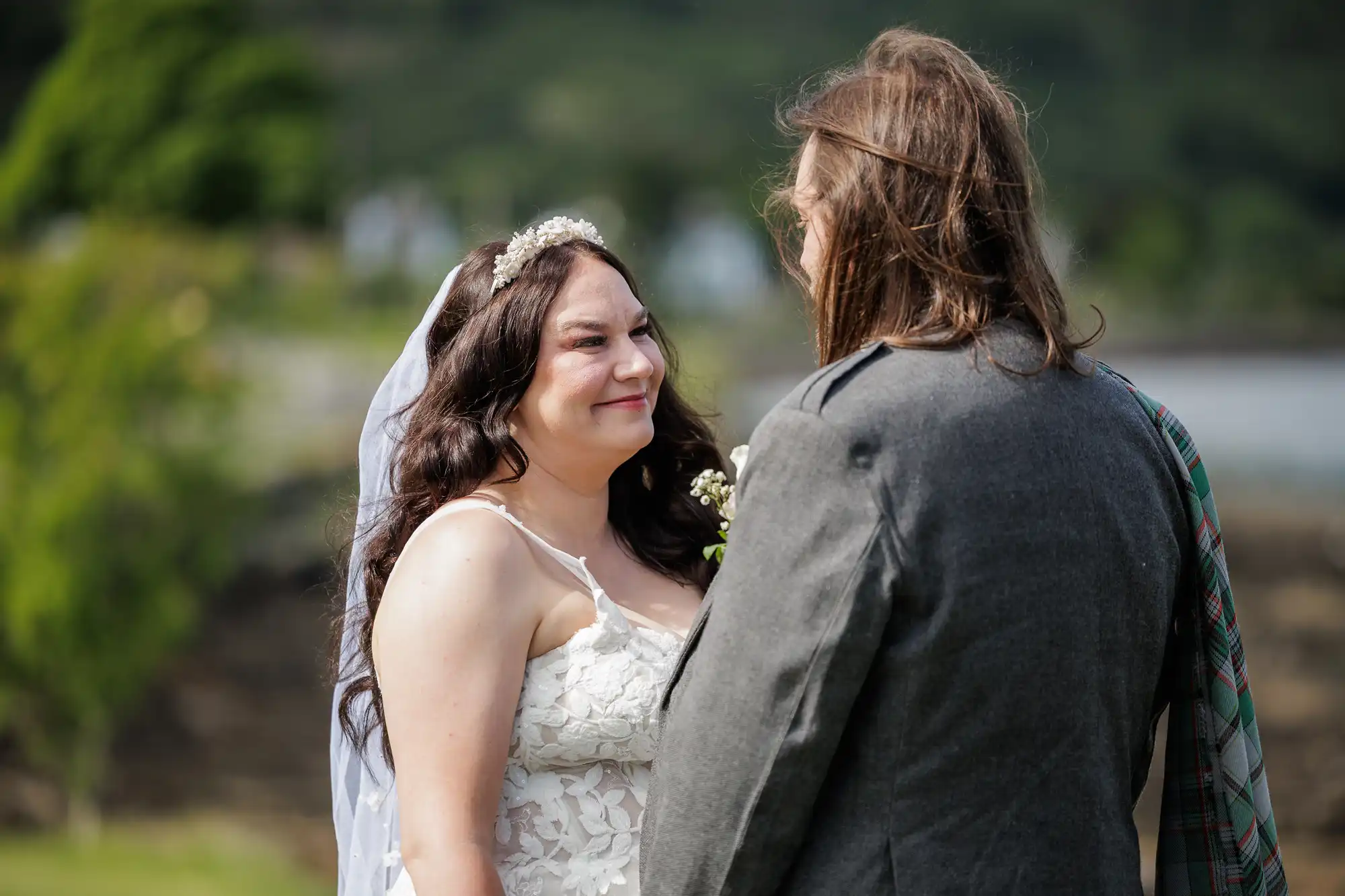 A bride in a white lace dress and veil smiles at a groom in a grey suit with long hair. They stand outdoors with greenery and blurred nature in the background, suggesting a wedding ceremony in a scenic, natural setting.
