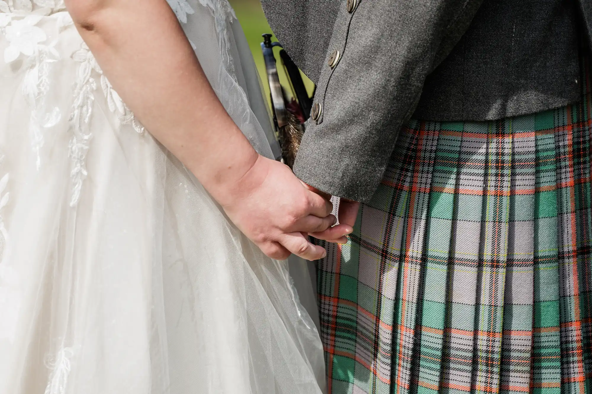 Close-up of a bride in a white lace dress and a groom in a grey jacket and green tartan kilt, holding hands. Only their torsos and hands are visible, highlighting their clasped hands in a tender, intimate moment.