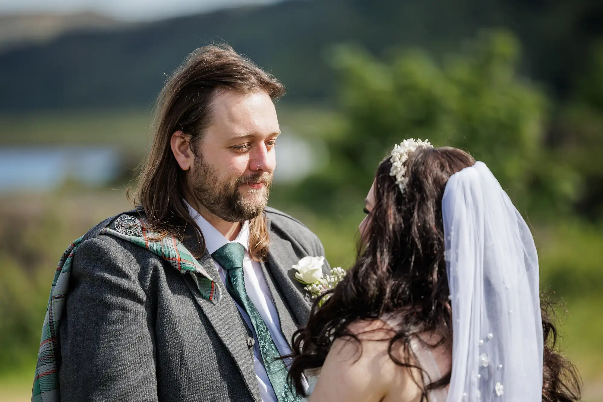 A groom in a grey suit with a tartan sash gazes at a bride in a white dress and veil adorned with flowers. They stand outdoors in sunlight, with greenery and a blurred lake and hills in the background, sharing an intimate moment.