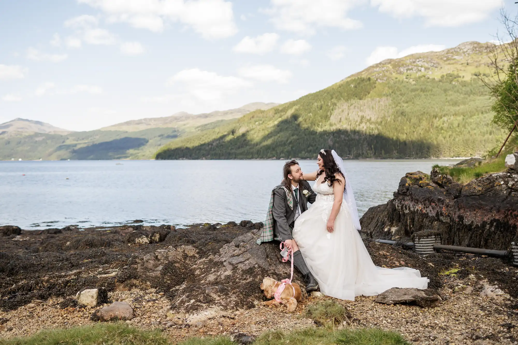 A bride and groom sit on rocky ground by a lake with green hills in the background. The groom, in a suit with a tartan kilt, kisses the bride’s hand. She wears a white dress and headband. A small brown dog with a pink bow sits in front of them.