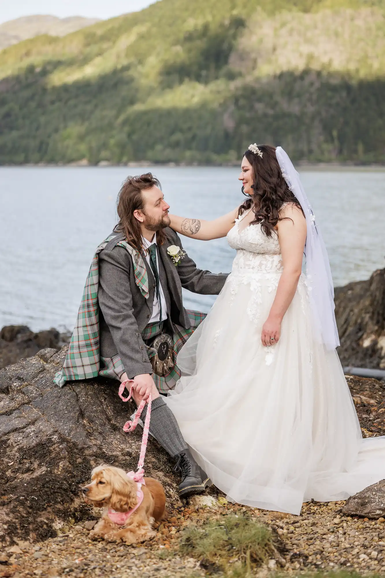 A bride in a white lace dress and veil stands on rocks beside a groom wearing a tartan kilt and grey jacket. He sits, holding her waist and a small brown dog on a pink lead. Behind them is a lake and green, forested mountains under a cloudy sky.