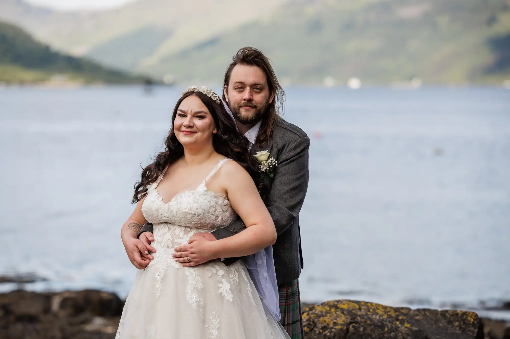 A bride in a white, lace wedding dress and a groom in a grey jacket with a kilt stand by a lake, embracing and smiling. Behind them, mountains and water stretch into the distance under a cloudy sky. Both look happy and relaxed.