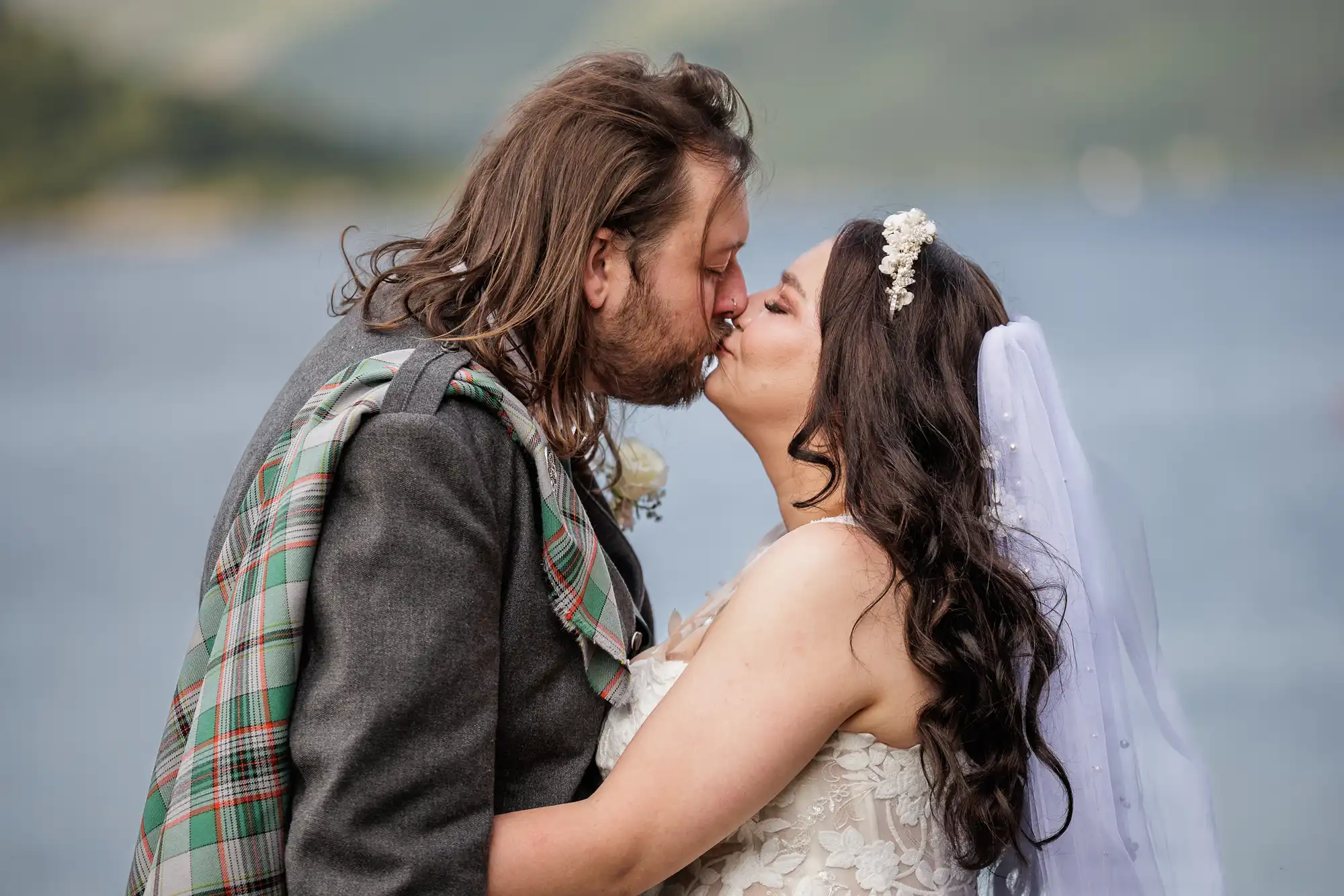 A bride and groom share a kiss outdoors. The groom wears a grey jacket with a tartan sash, and the bride wears a white lace dress with a veil and floral headpiece. A lake and green hills are softly blurred in the background.