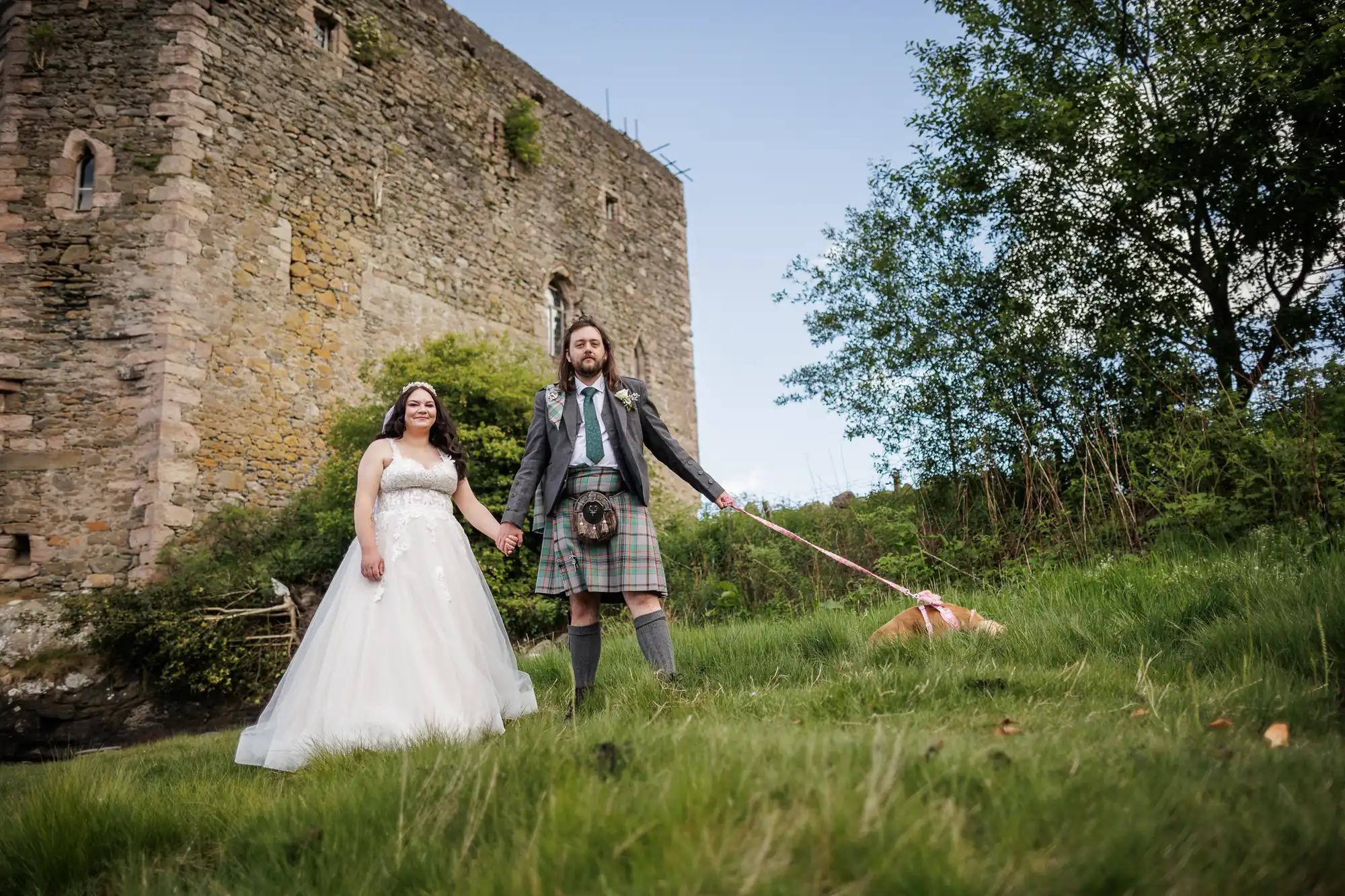 A bride in a white dress and a groom in a kilt stand holding hands on grass near a stone castle. The groom holds a lead attached to a dog that is sniffing the ground. Trees and greenery surround the couple and castle.