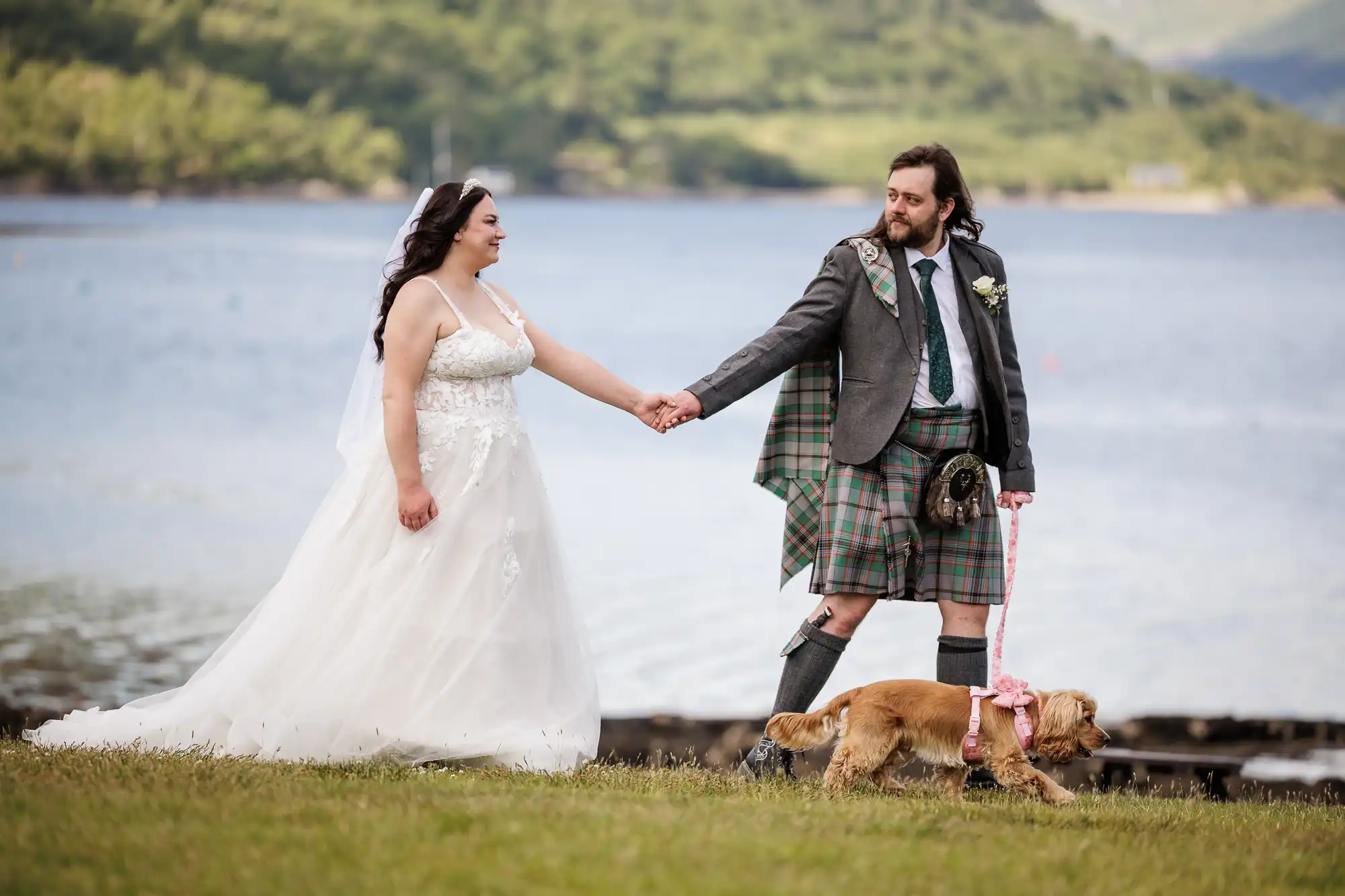 A bride in a white gown and a groom in a Scottish kilt hold hands while walking outdoors by a lake. The groom holds a lead, walking a small brown dog. Green hills and water are visible in the background, creating a serene scene.