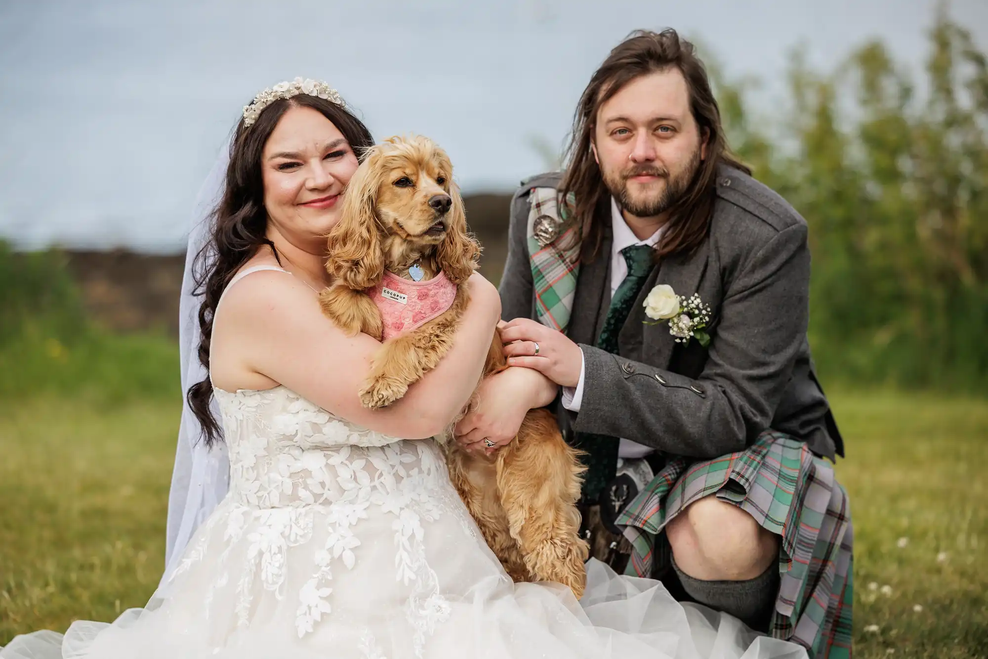 A bride in a white lace wedding dress holds a brown cocker spaniel wearing a pink harness. Next to her, a groom in a grey jacket and tartan kilt kneels, smiling. They are outdoors on grass with greenery and a cloudy sky in the background.