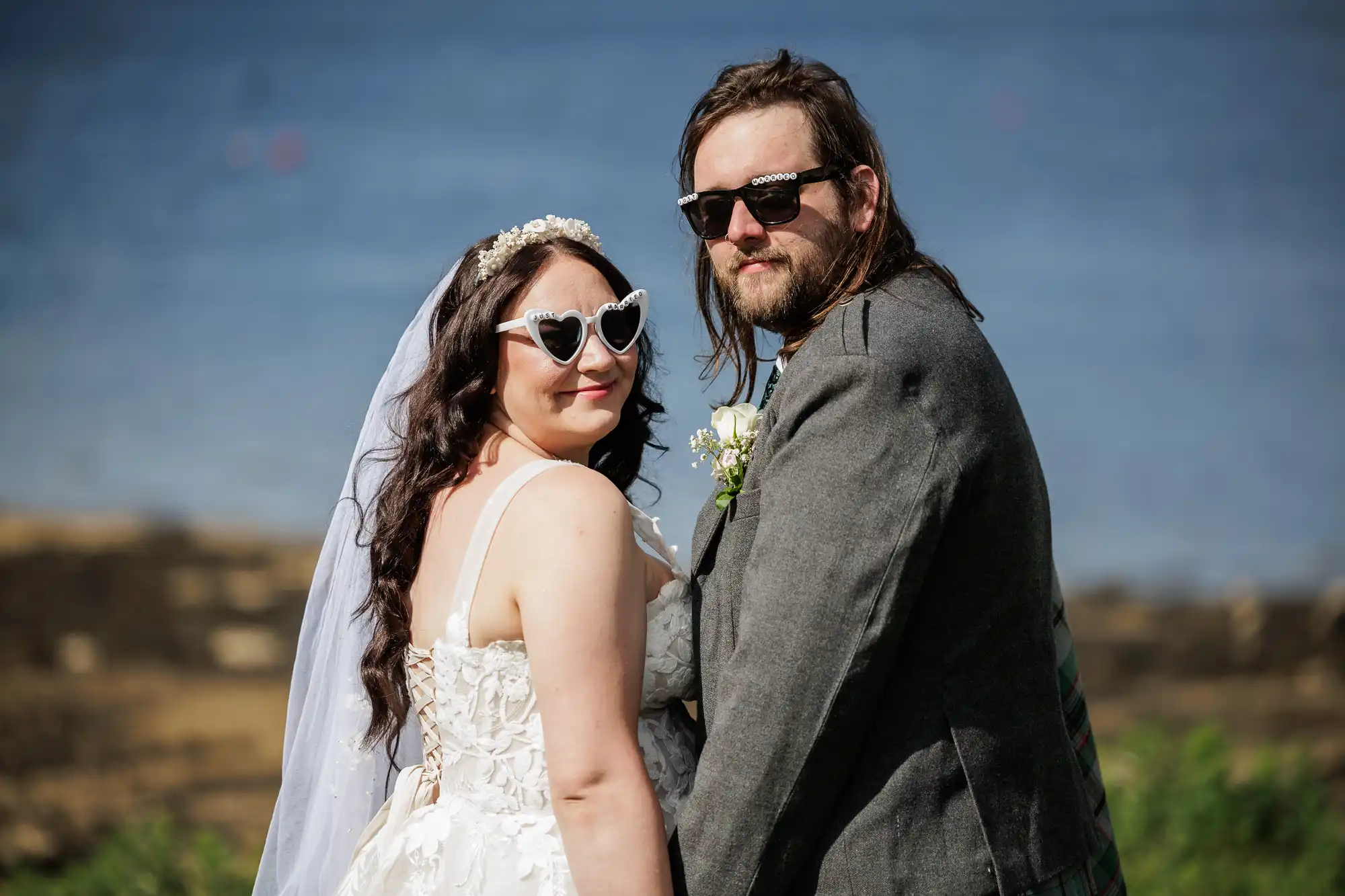 A bride in a white dress and heart-shaped sunglasses stands beside a groom in a grey suit and black sunglasses. They hold hands, smiling at the camera, with the sea and rocky shore blurred in the background.