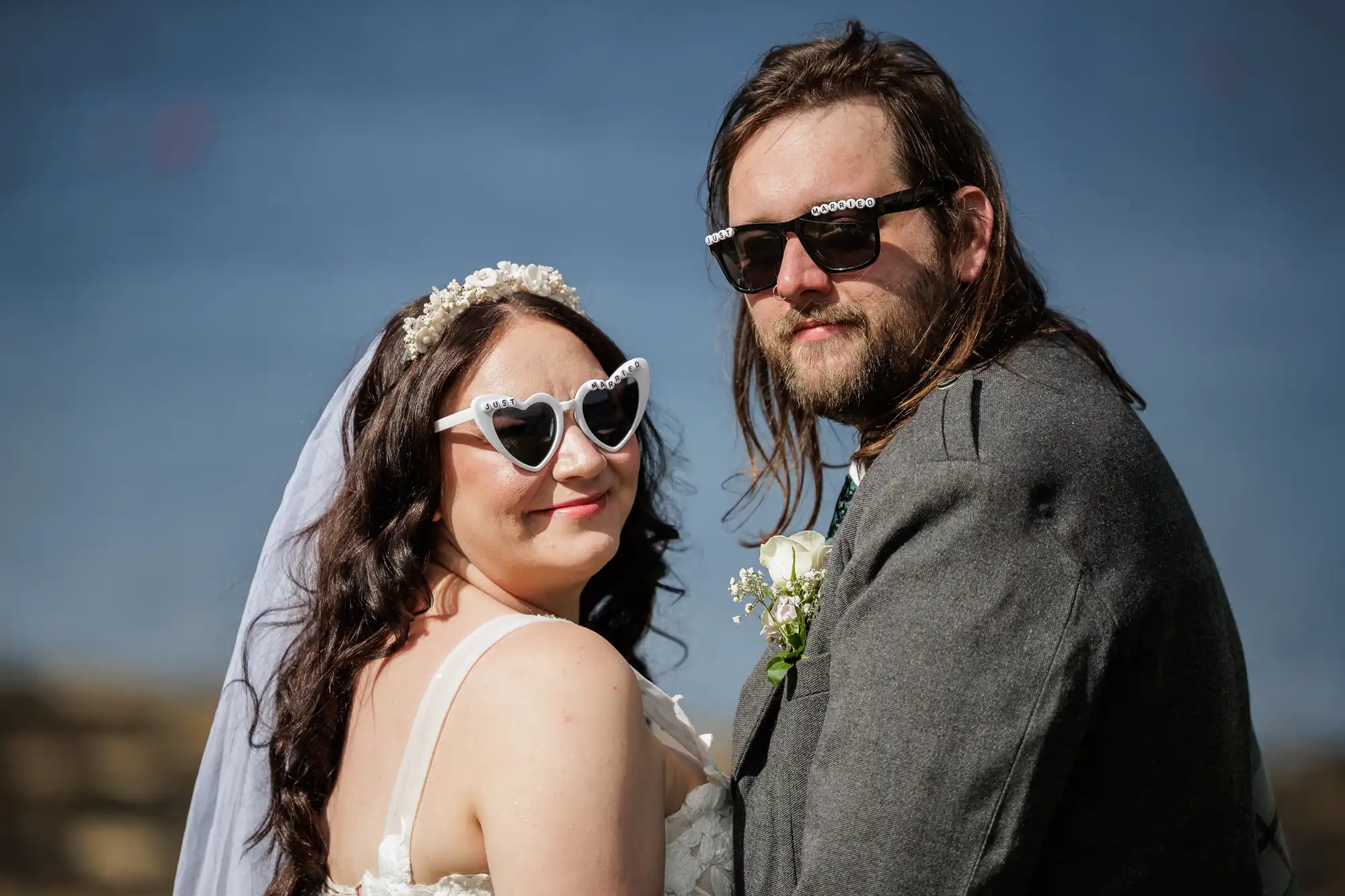 A bride and groom stand close together outdoors, both wearing heart-shaped sunglasses. The bride wears a white dress, veil, and flower crown; the groom wears a grey suit with a buttonhole. They smile, looking over their shoulders at the camera. Blue sky background.